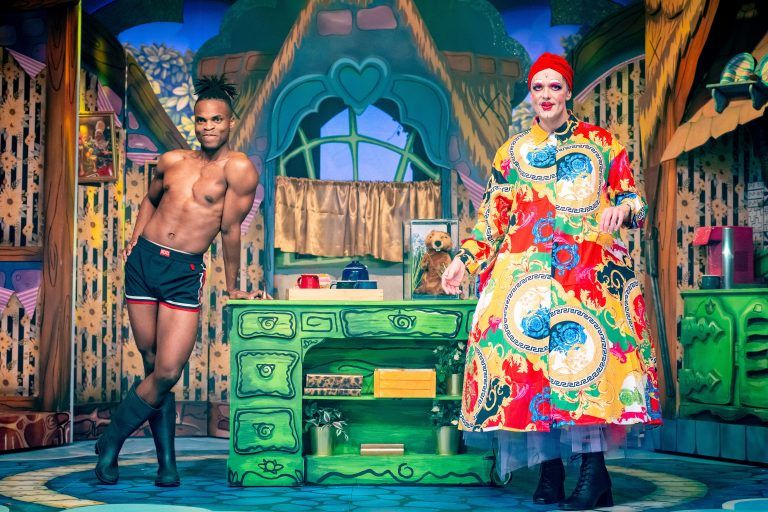 Well toned black man in his 20s poses in his underwear in a pantomime kitchen, next to a white panto dame wearing an outlandishly large housecoat of many colours.