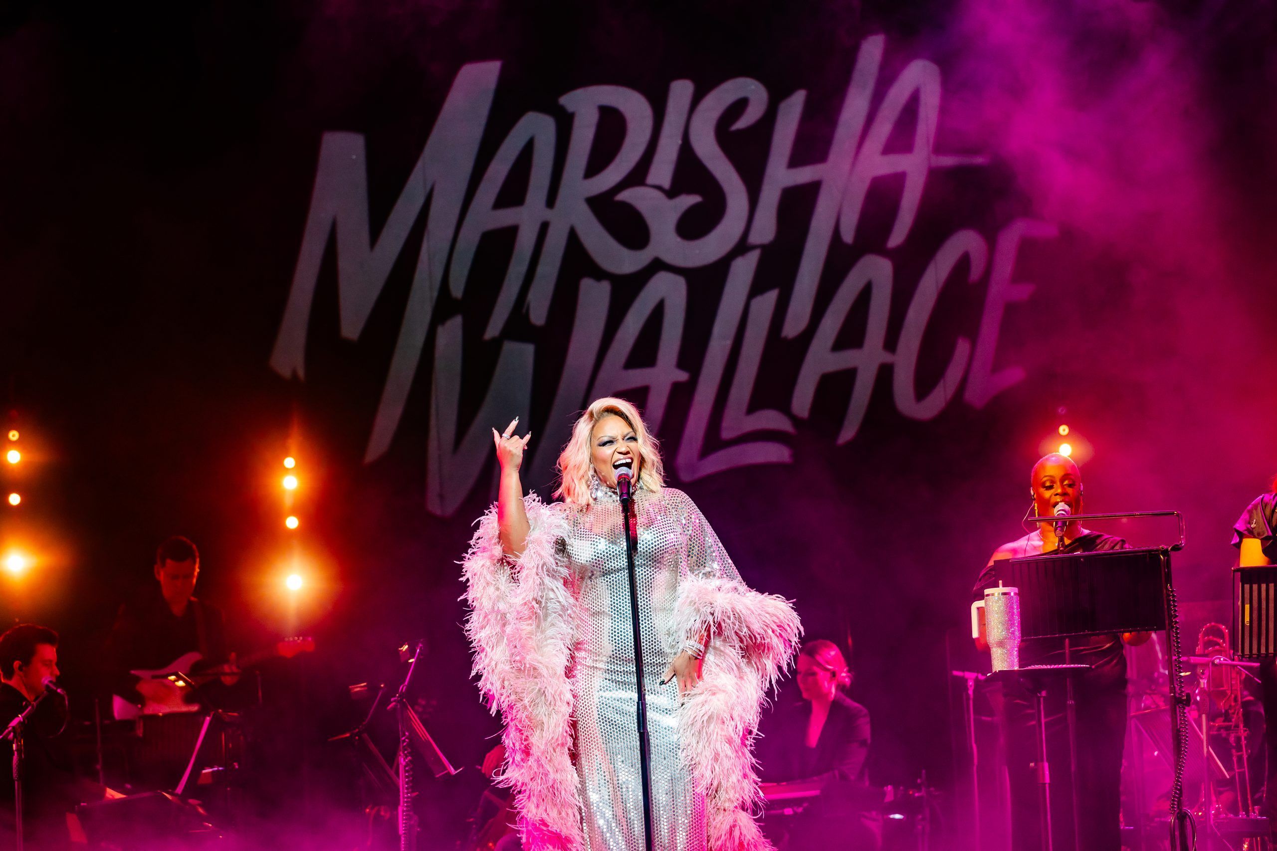 With "Marisha Wallace" projected behind her, she sings in a dazzling silver gown, arms wide in front of glowing stage lights.