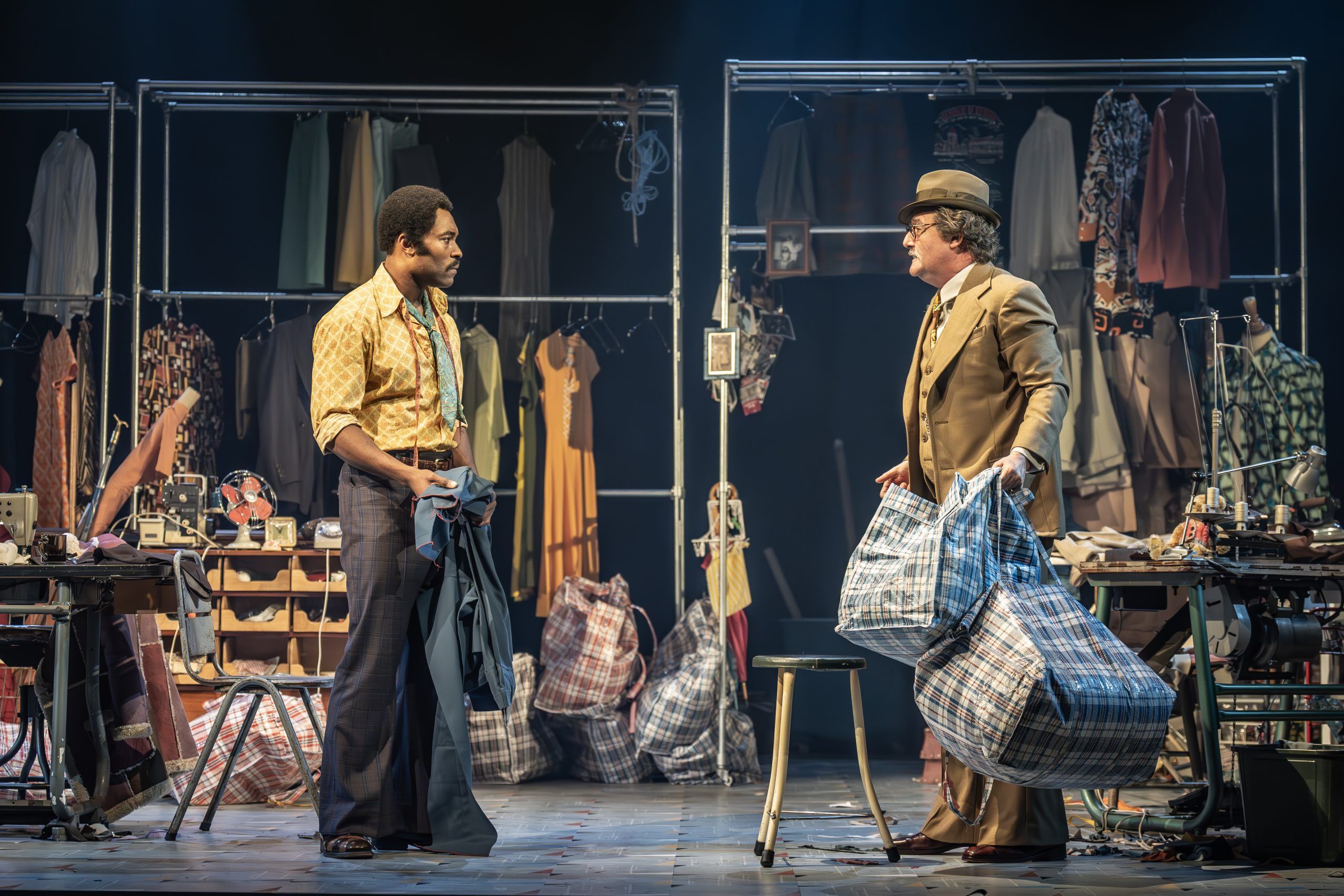 Alt text: Arinzé Kene as Walker Holt and Colin Mace as Mr Nat face each other on stage in a tailor shop, surrounded by piles of checked laundry bags and sewing machines. Mr Nat holds two full bags while Walker listens, holding garments.