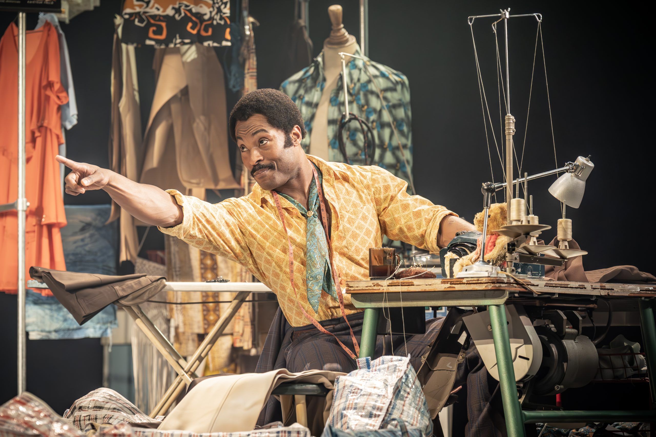 Arinzé Kene as Walker Holt sits at a sewing machine, mid-gesture and pointing, with a playful expression. He’s surrounded by fabric and tools in a busy tailor shop set.