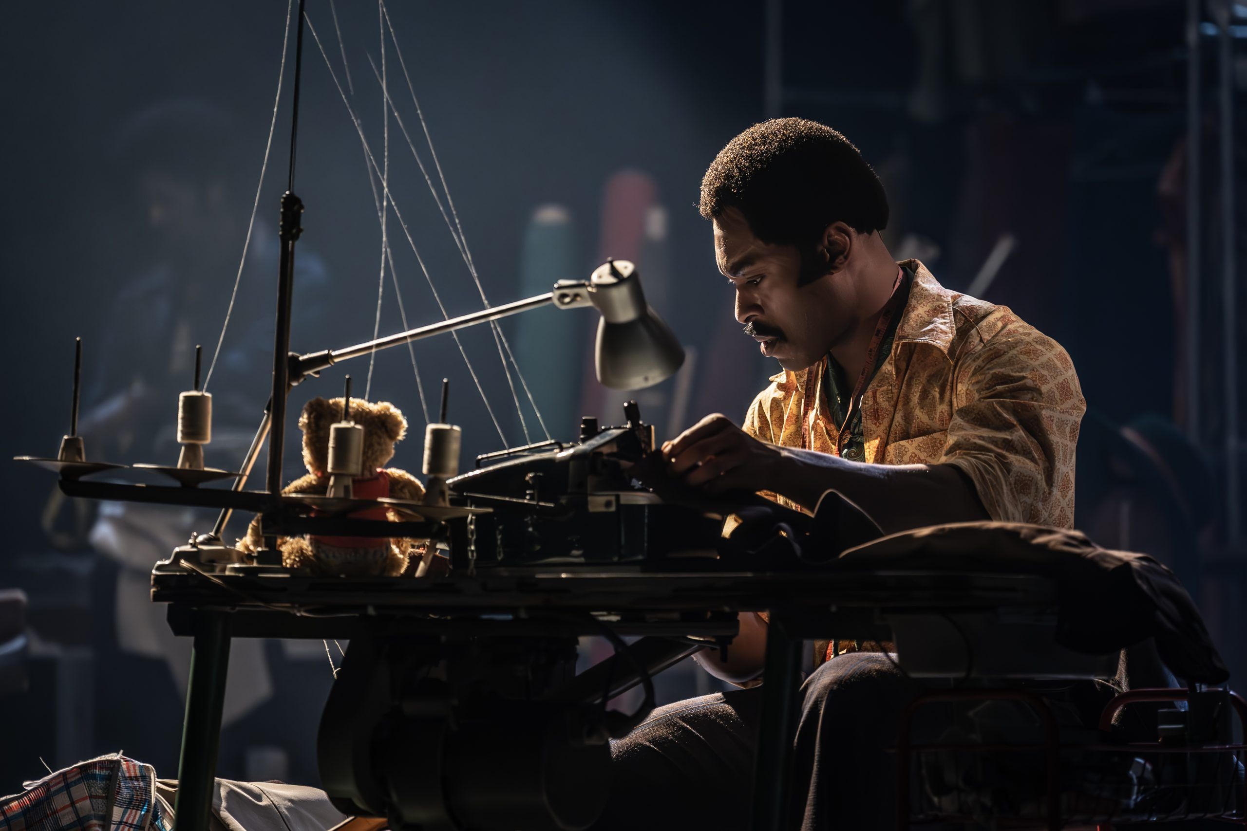 Arinzé Kene as Walker Holt sits at a sewing machine, focused and lit by a single desk lamp. A small teddy bear is perched above the machine, surrounded by thread spools and fabric in a dimly lit tailor shop.