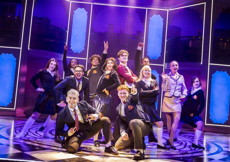 The cast of Cruel Intentions: The '90s Musical UK and Ireland Tour 2025 strike a confident and playful pose in a high-energy ensemble moment. Dressed in preppy school uniforms with plaid skirts, blazers, and ties, they embody the elite Manhattan high school setting of the show. At the center, Nic Myers as Kathryn stands with hands on her hips, while Will Callan as Sebastian exudes smug confidence in his burgundy blazer. The neon-lit set, featuring stylised window panels and a New York skyline backdrop, adds a sleek, modern touch. (Photo credit: Pamela Raith Photography)