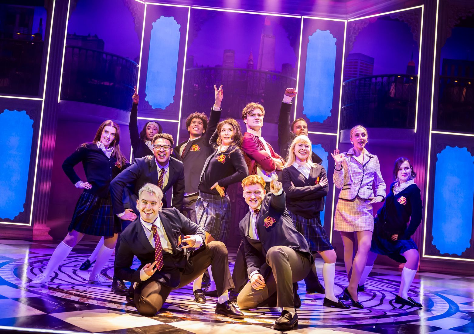 The cast of Cruel Intentions: The '90s Musical UK and Ireland Tour 2025 strike a confident and playful pose in a high-energy ensemble moment. Dressed in preppy school uniforms with plaid skirts, blazers, and ties, they embody the elite Manhattan high school setting of the show. At the center, Nic Myers as Kathryn stands with hands on her hips, while Will Callan as Sebastian exudes smug confidence in his burgundy blazer. The neon-lit set, featuring stylised window panels and a New York skyline backdrop, adds a sleek, modern touch. (Photo credit: Pamela Raith Photography)