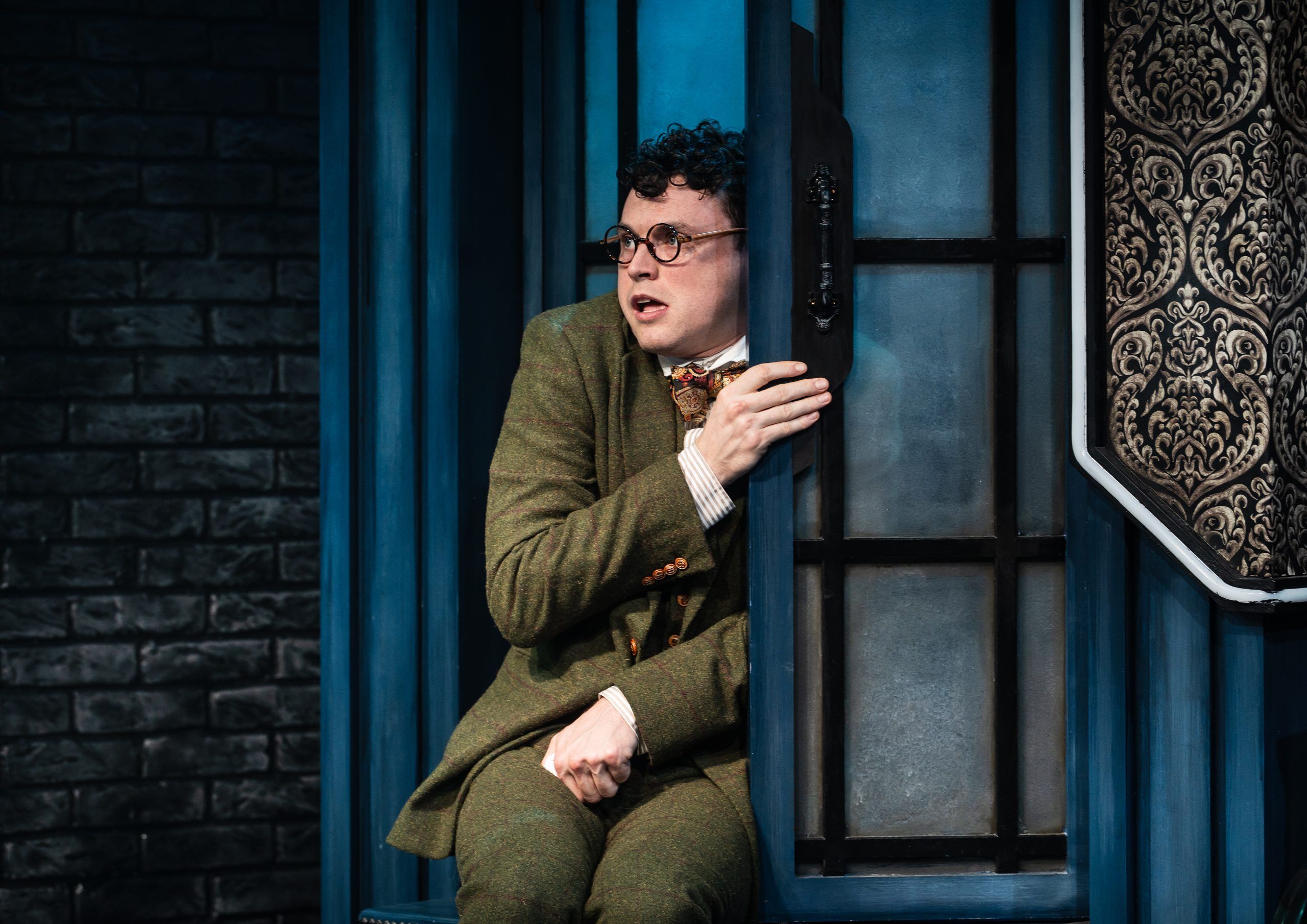 A curly-haired man in round glasses and a green three-piece suit clutches a doorframe, wide-eyed and worried, against a blue-lit gothic backdrop.