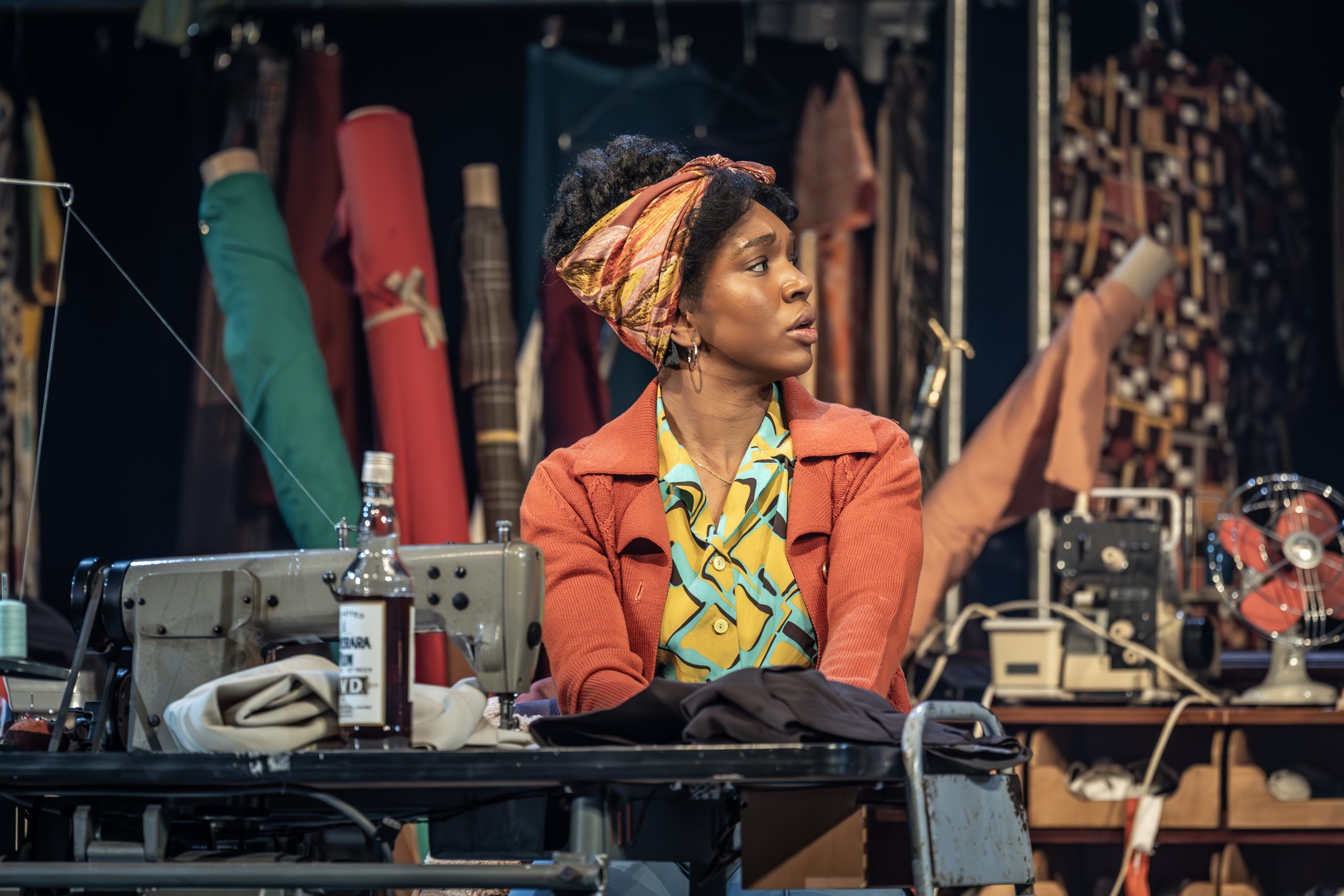 Cherrelle Skeete as Darlene Holt sits at a sewing machine, looking to her right with a concerned expression. She wears a colourful headscarf and 70s outfit, surrounded by fabric and tools.