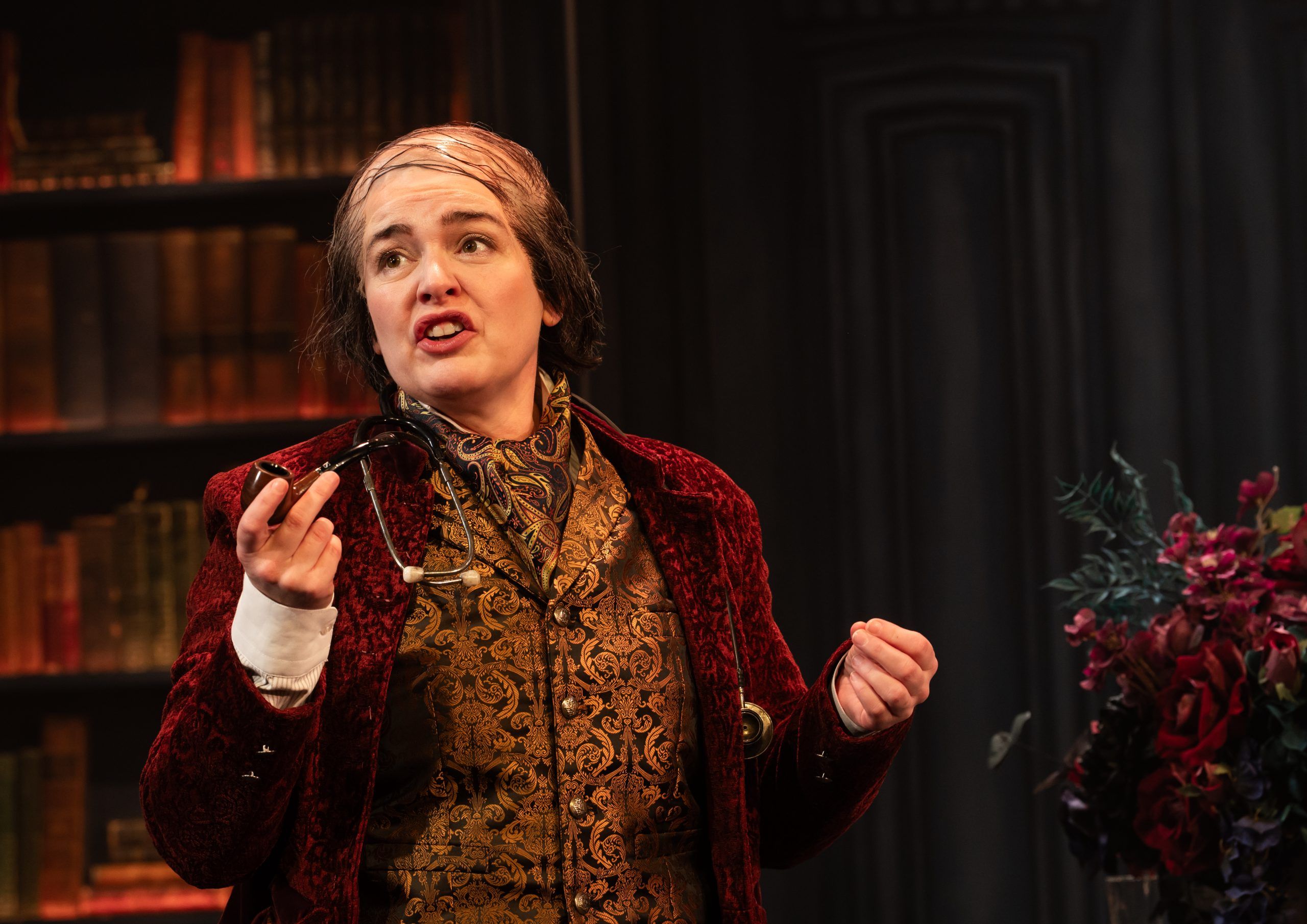 A woman playing a man in a red velvet coat, greying wig and cravat, holding a pipe and stethoscope, speaks animatedly in front of a library backdrop.