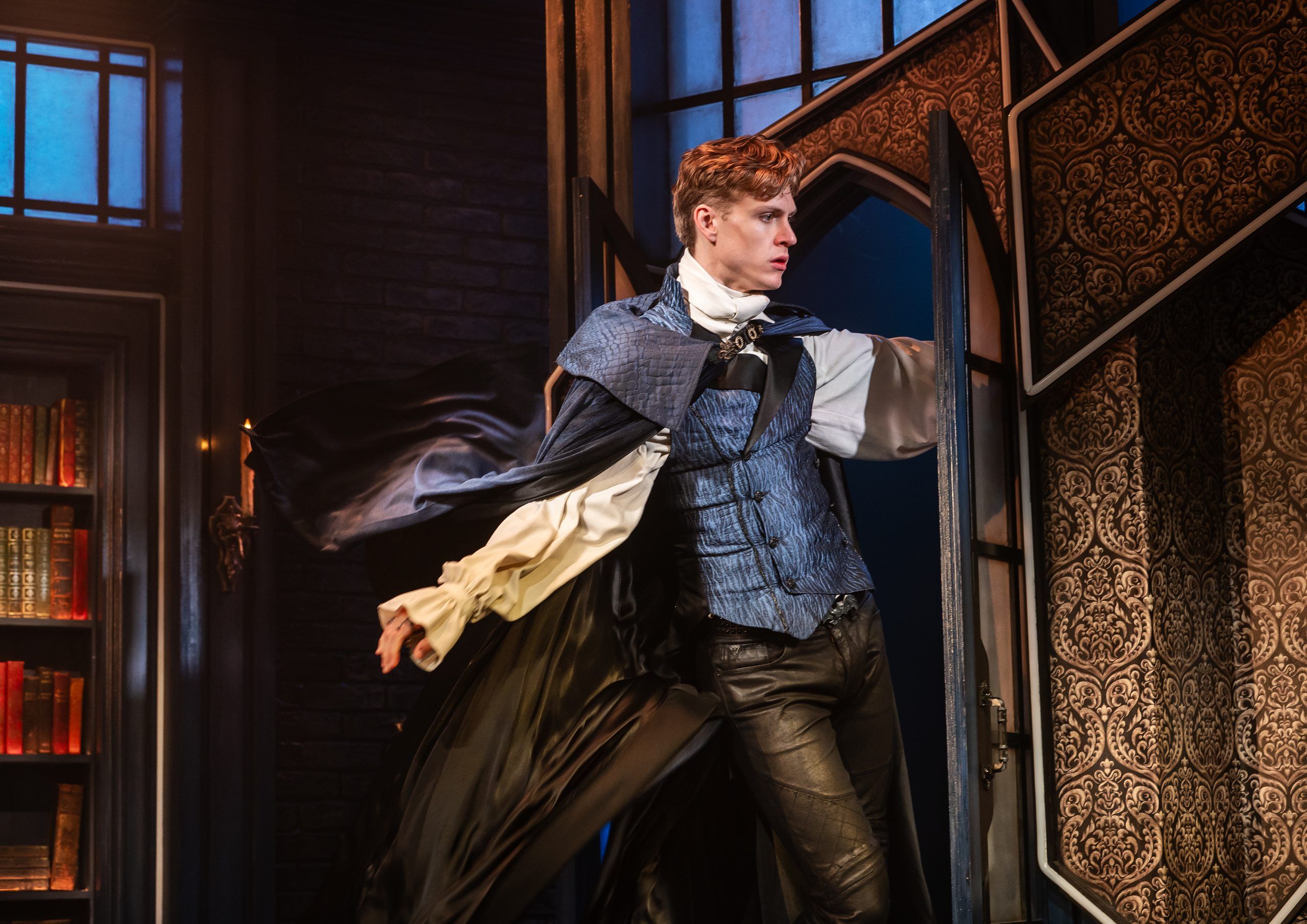 Dracula bursts through a gothic window, cape billowing, his expression determined, in a fitted blue waistcoat and puffy white shirt.