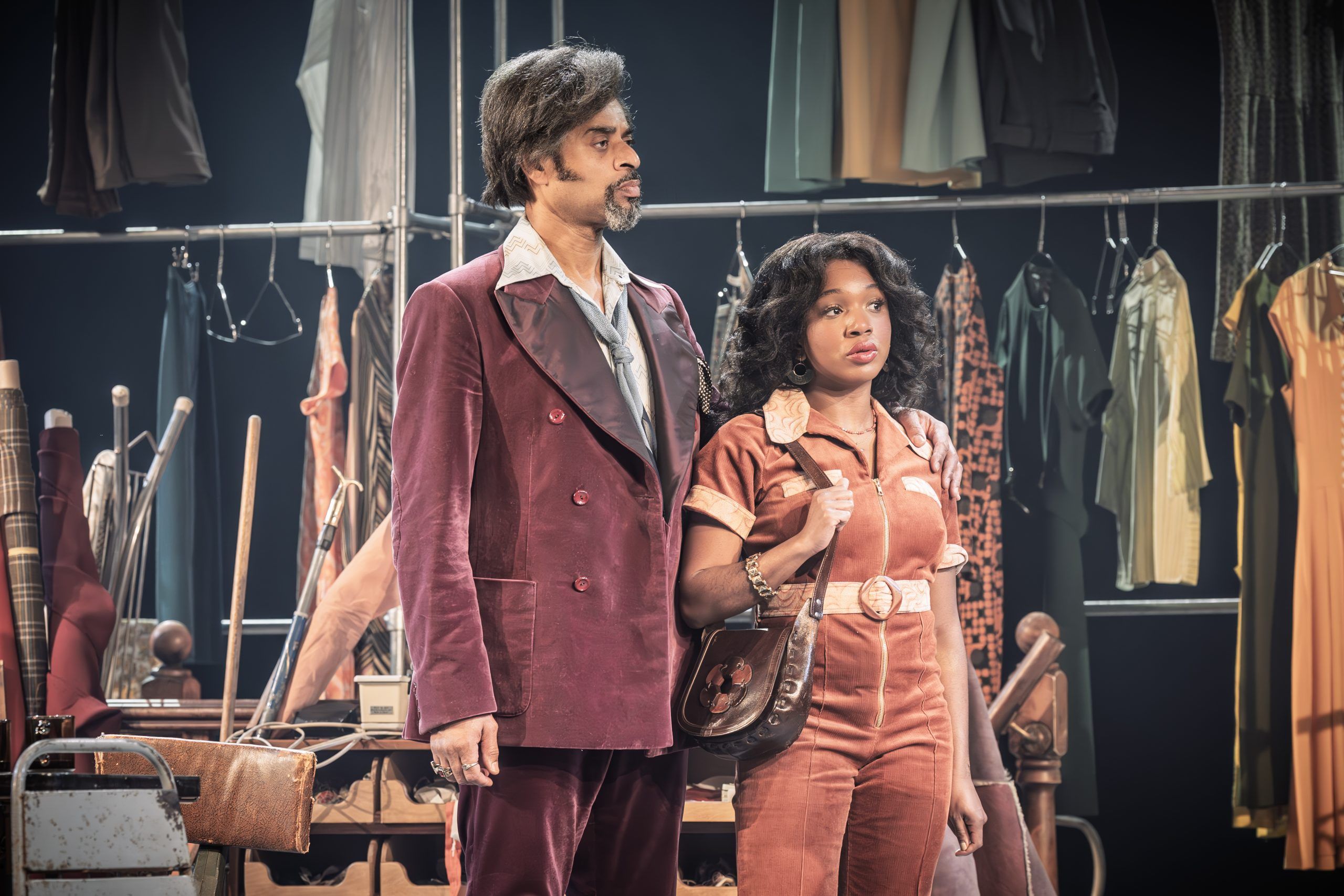 Karl Collins as Horace stands beside Cherrelle Skeete as Darlene in a matching rust-coloured jumpsuit. They both look ahead with solemn expressions, framed by 70s garments on rails behind them.