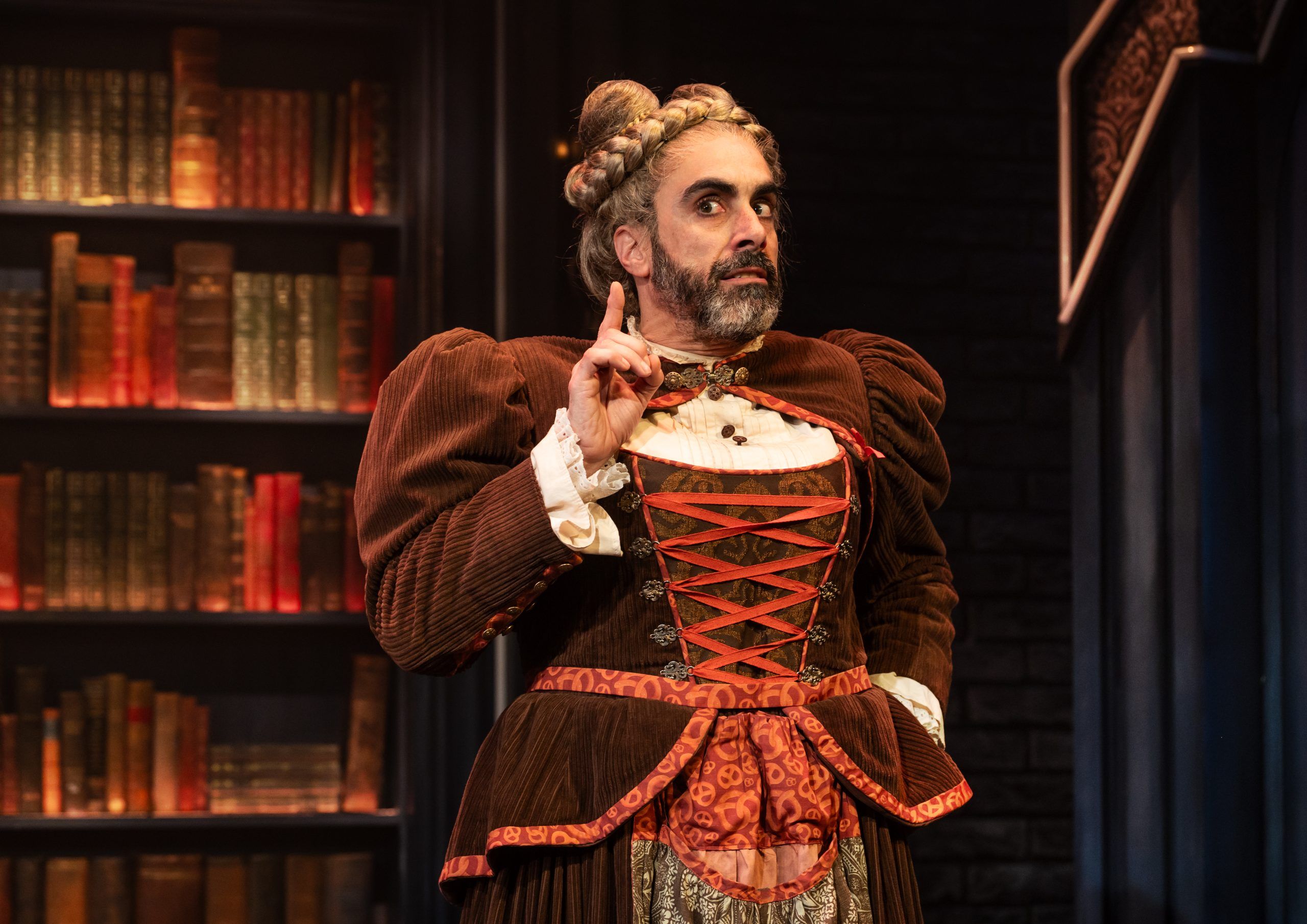 A bearded person in a braided wig and corseted brown gown gestures expressively with a raised finger, standing in front of ornate bookcases.