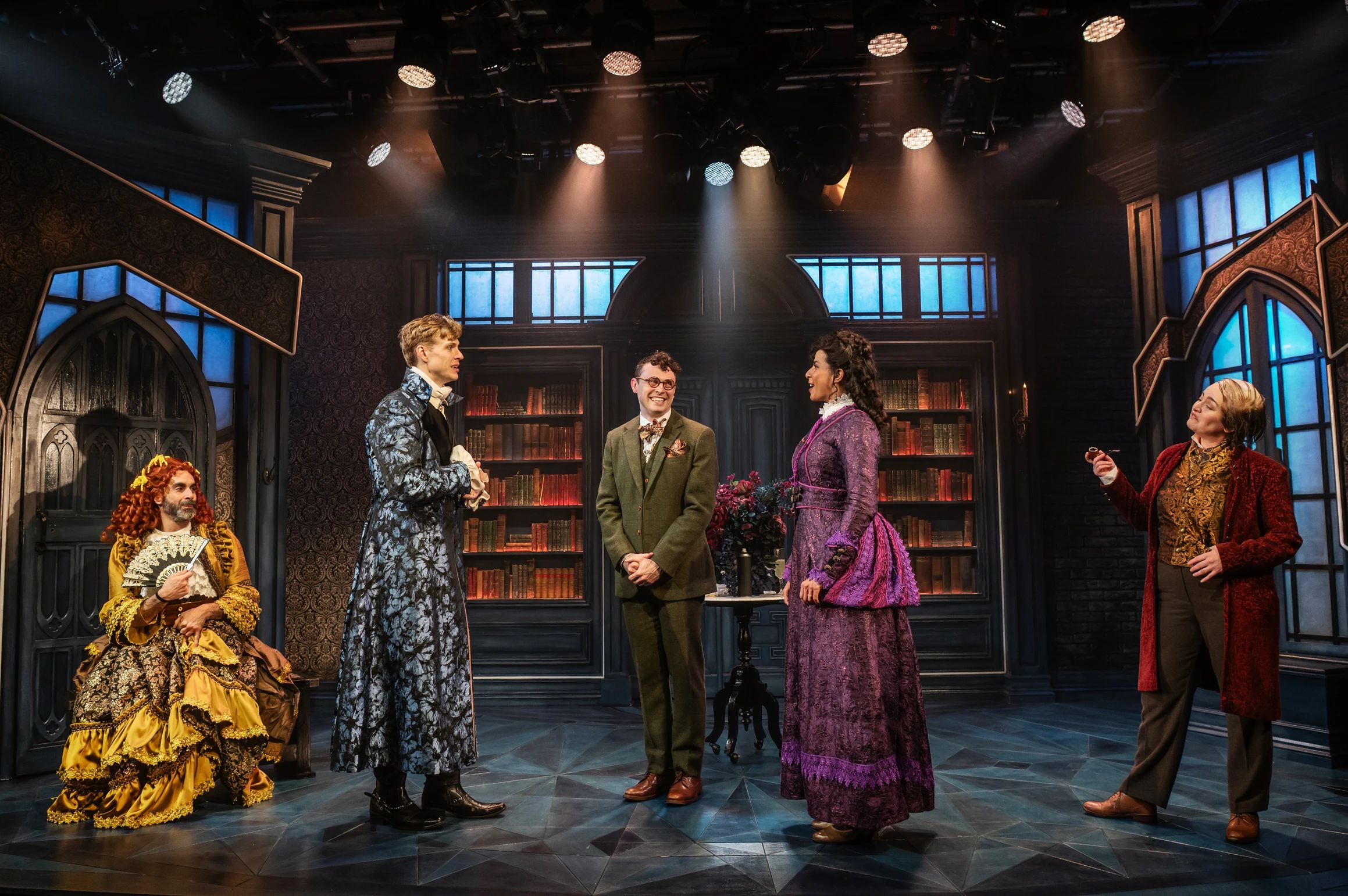 Five characters, all in elaborate period costumes, stand under spotlights in an ornate, book-lined room; one sits in an extravagant yellow gown with fan.