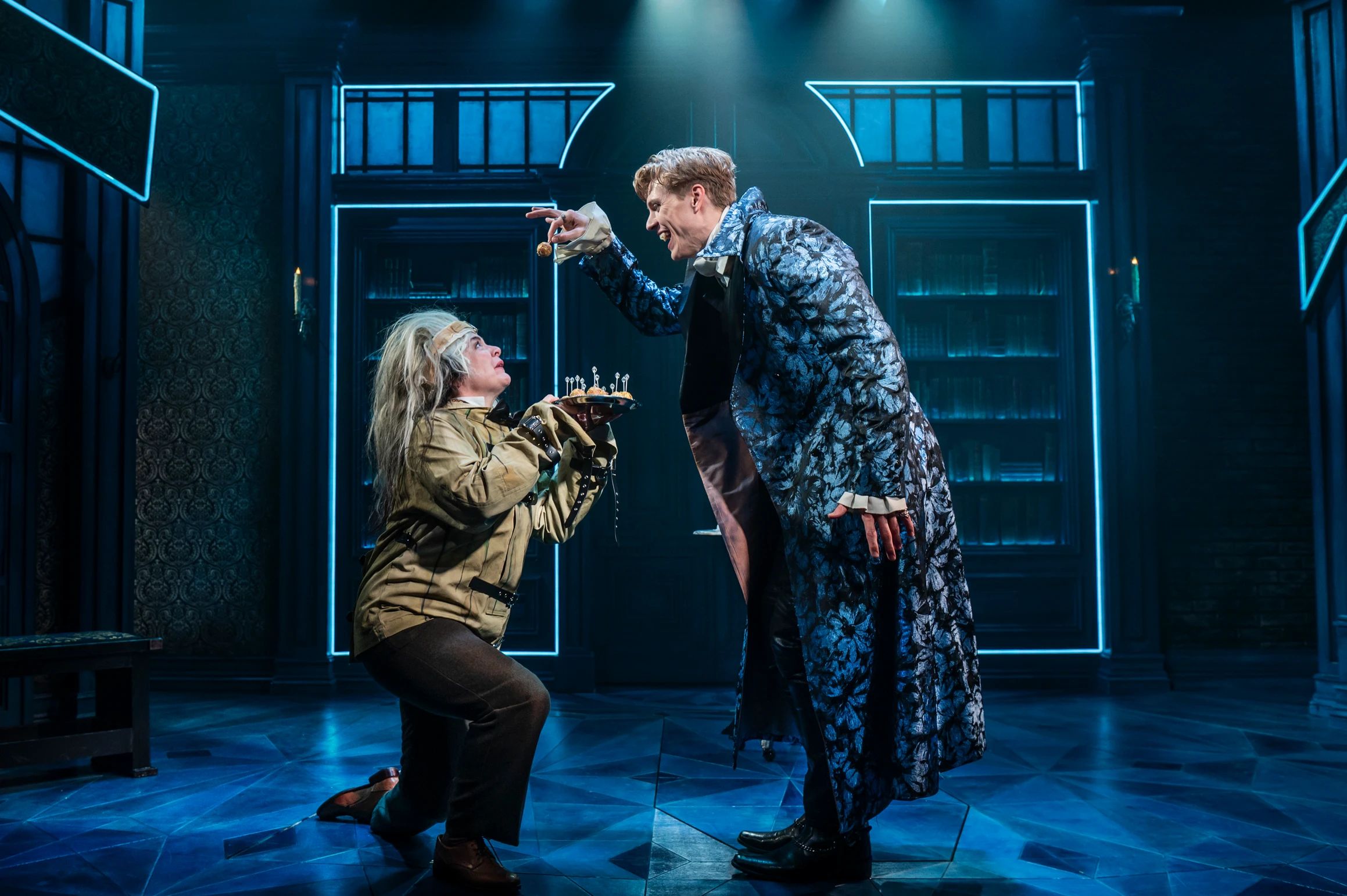 Dracula holds a treat just out of reach of a wild-haired Renfield, who kneels and offers a spiked cake on a tray, under moody blue lighting.