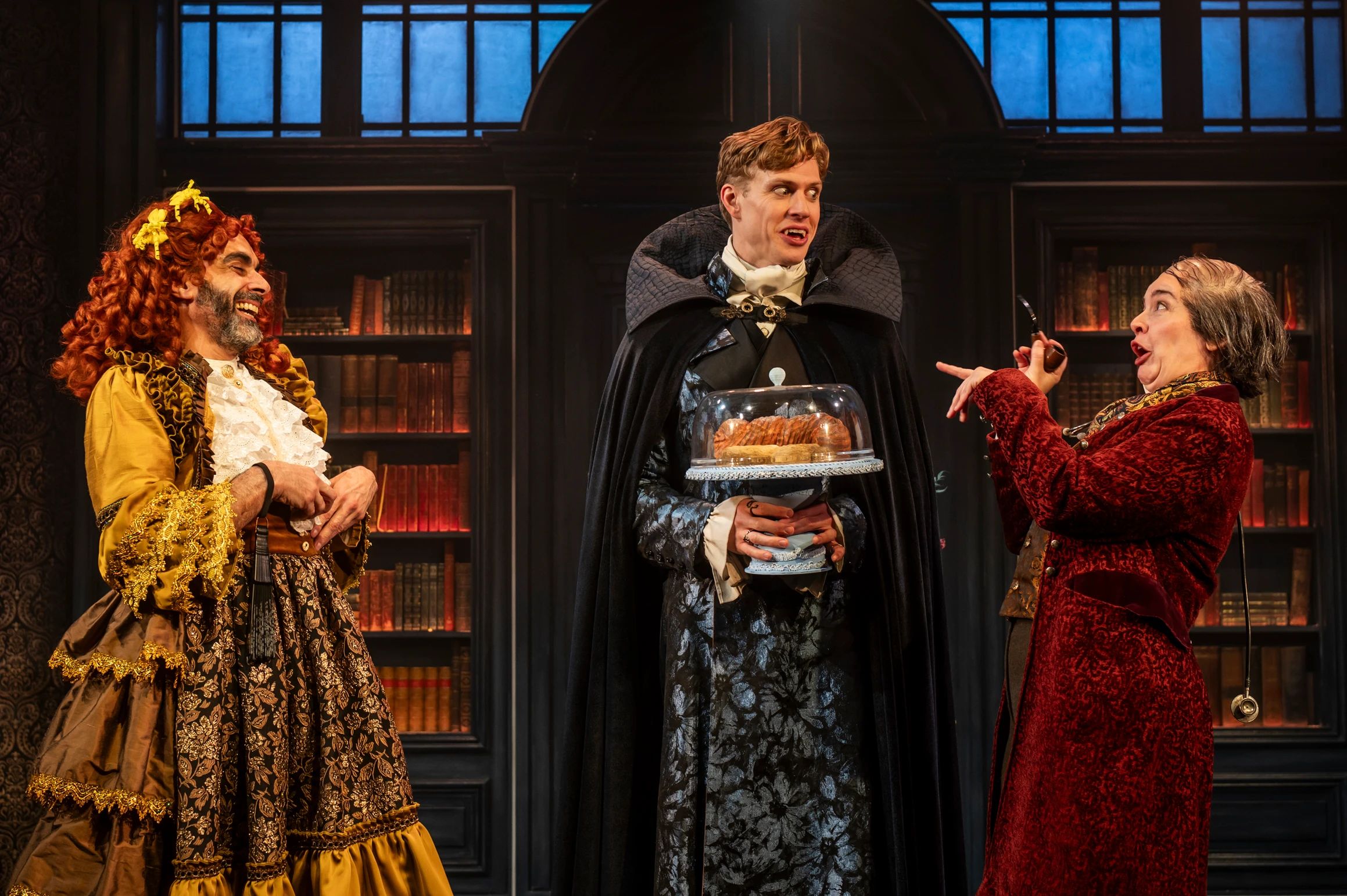 Dracula stands proudly with a domed cake tray while a flamboyant character in a yellow dress and another in red velvet react with exaggerated expressions.
