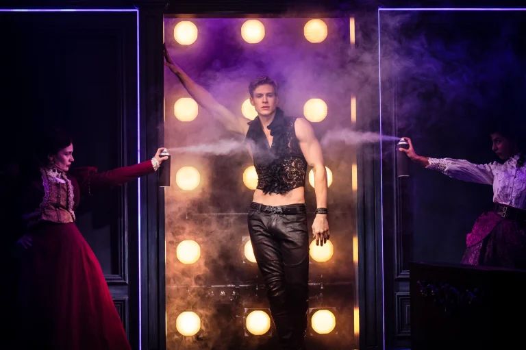 Dracula emerges through a mist-filled doorway in tight black trousers and a lacy black crop top, flanked by two women spraying fog.