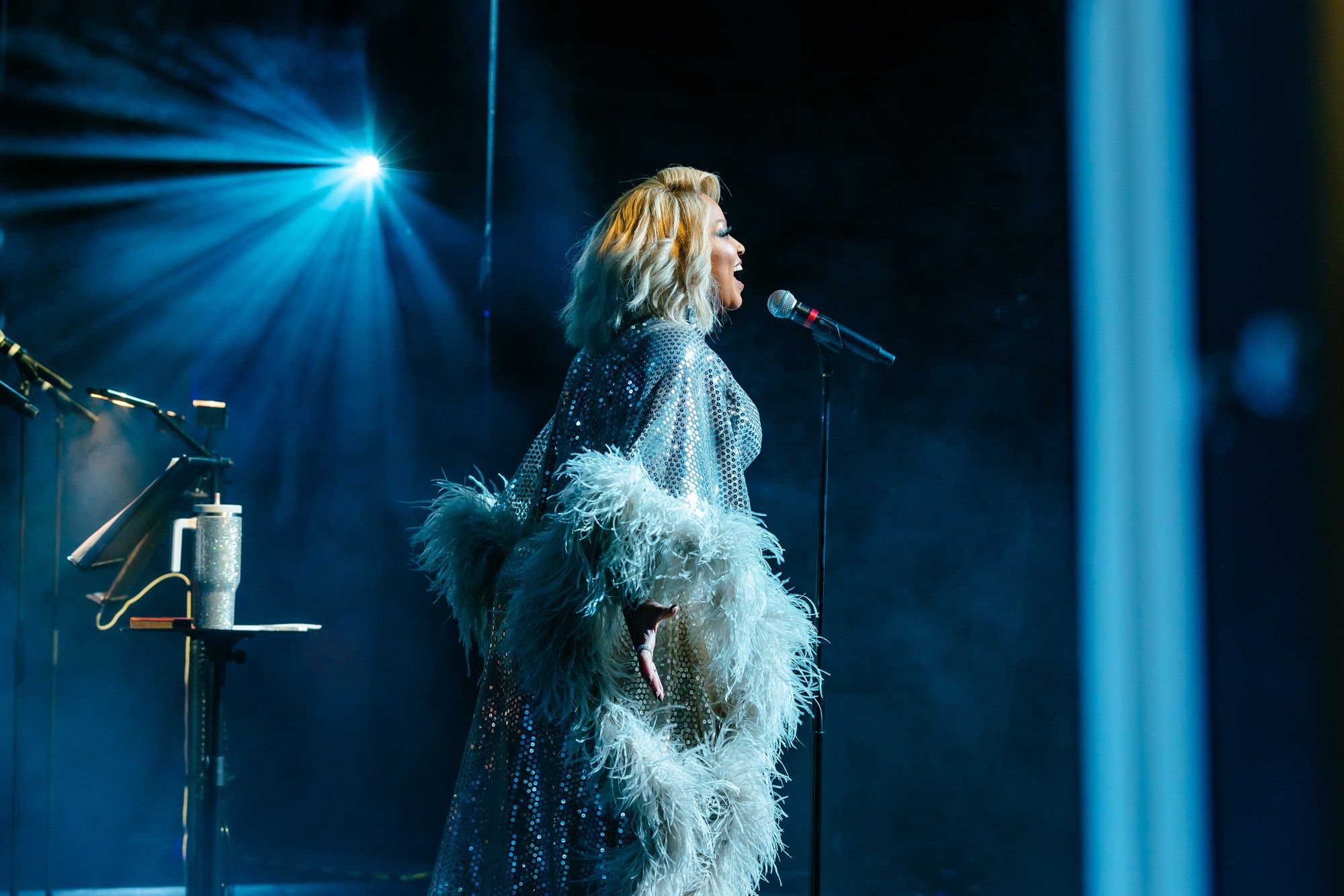 Performing in a silver sequined cape with feathered cuffs, Marisha Wallace belts out a note under piercing blue stage lights.