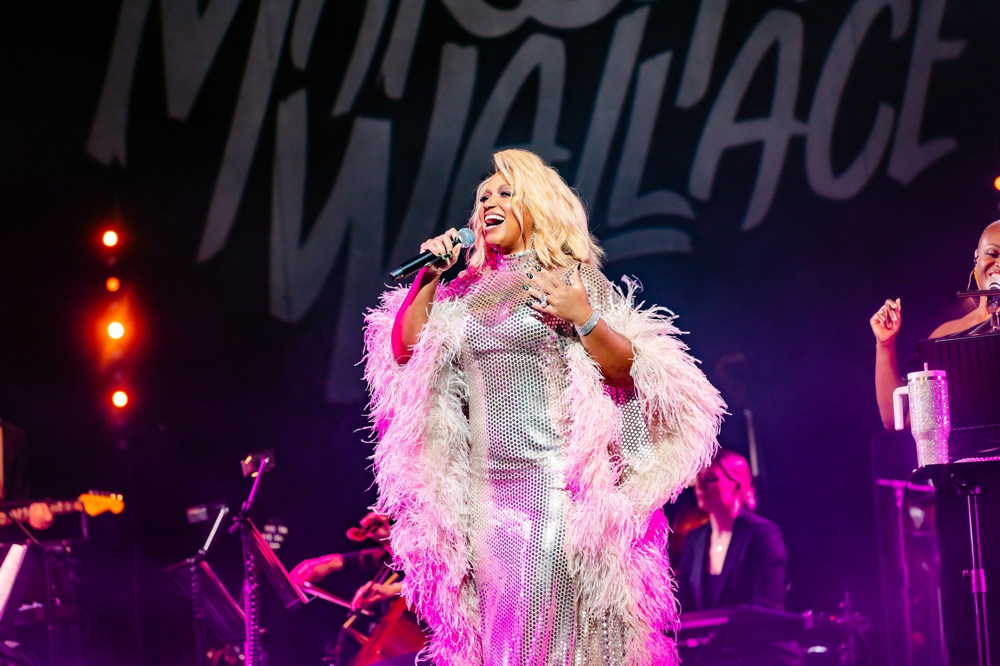 Smiling and holding the microphone, Marisha Wallace engages the audience in a shimmering silver gown with feathered trim.