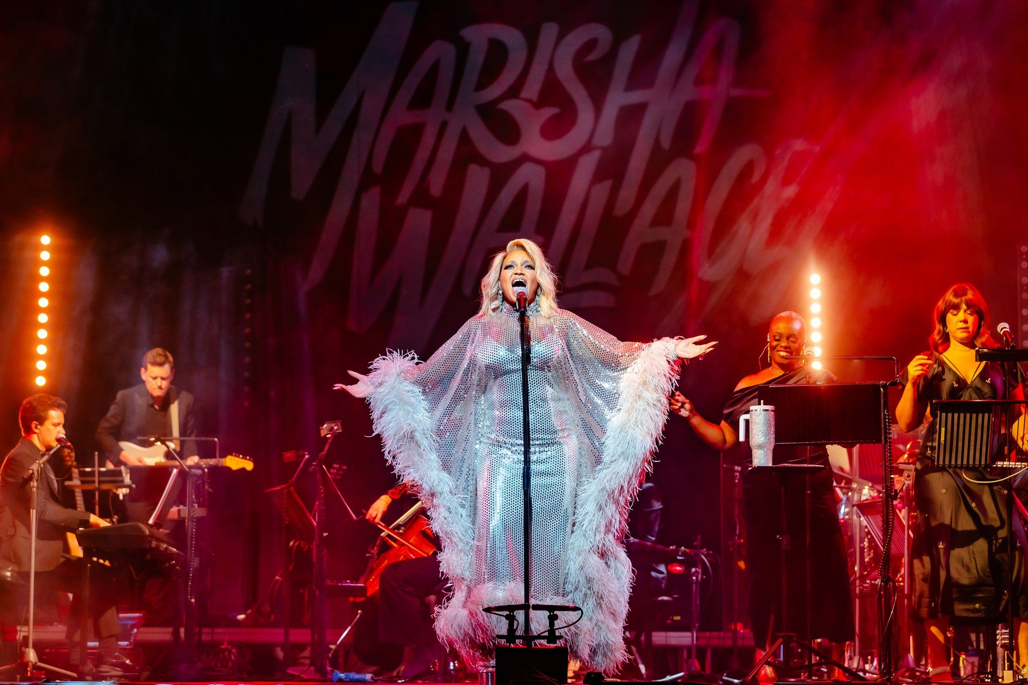 Marisha Wallace stands centre stage in a radiant silver gown, arms extended, flanked by her band and backup singers.