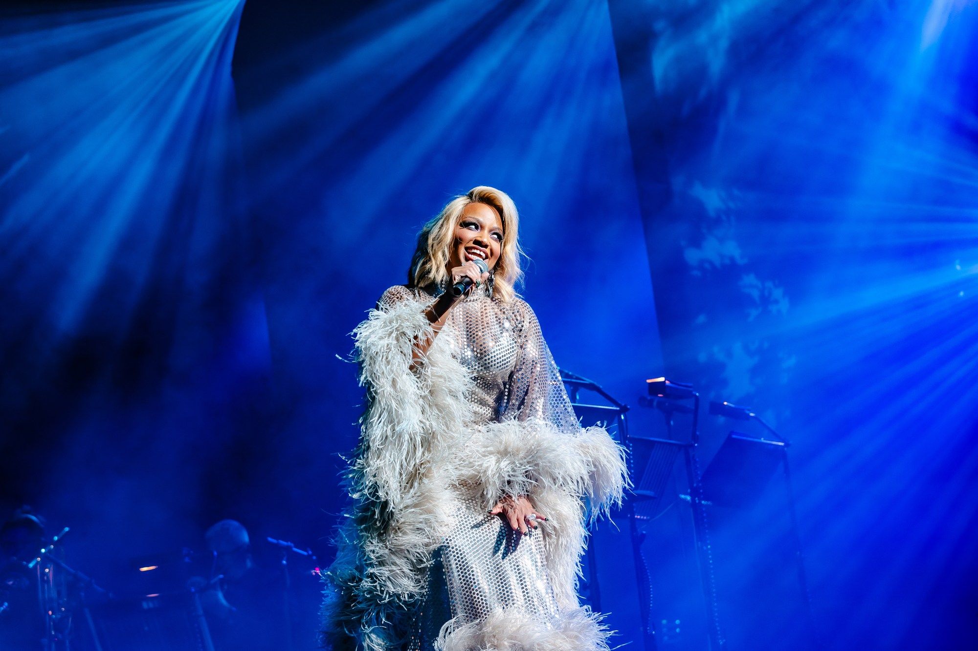 Bathed in blue light, Marisha Wallace beams while performing in a sequined gown with feathered sleeves, exuding confidence.