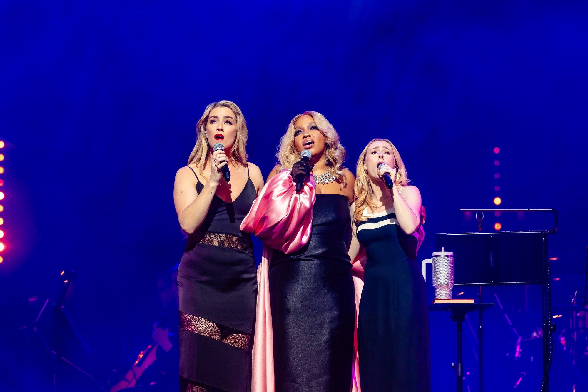 Marisha Wallace harmonises with two other singers, dressed elegantly in black, under rich blue stage lighting.