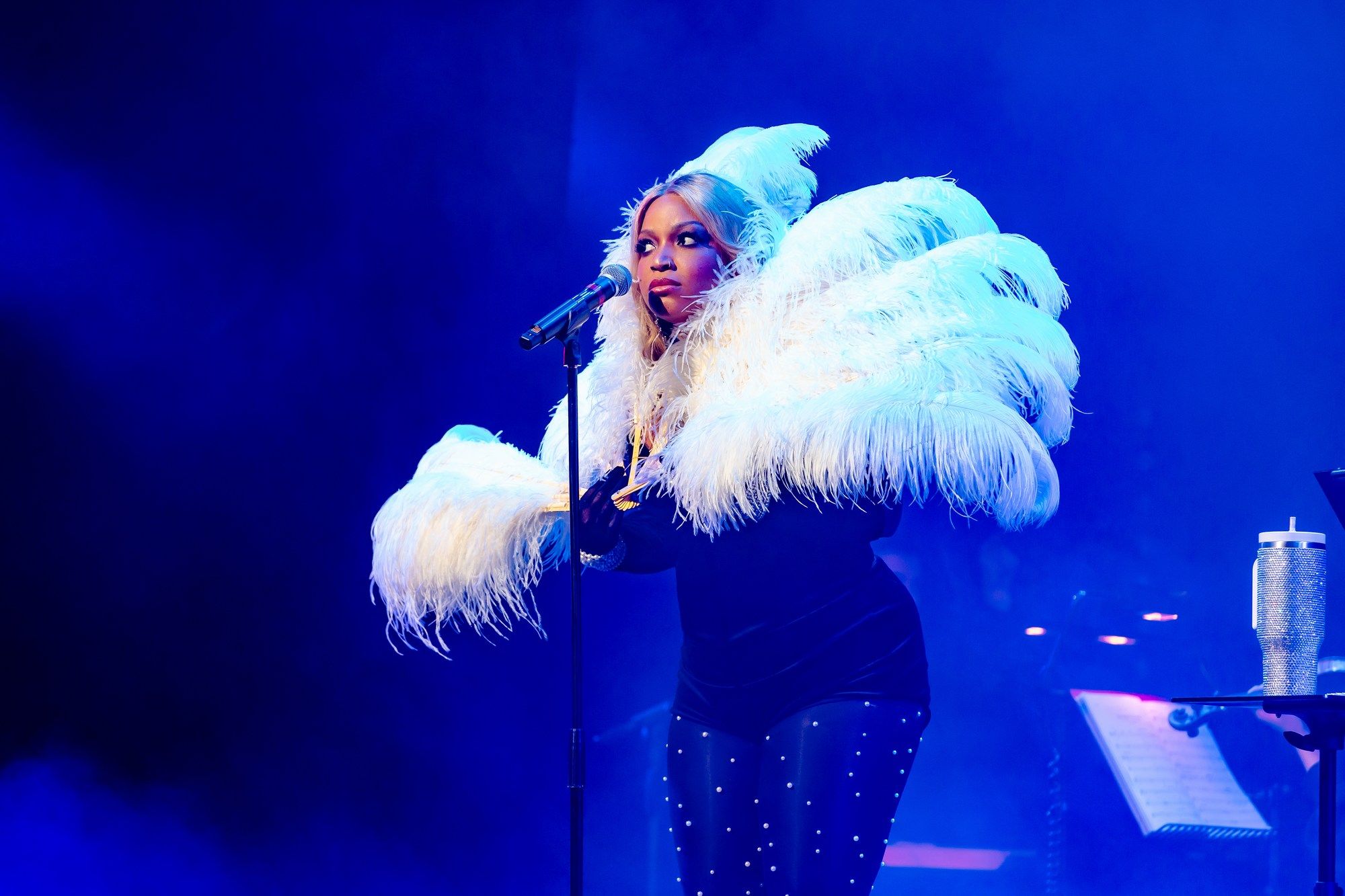 Marisha Wallace in a black outfit with dramatic white feathered sleeves leans into the microphone, bathed in moody blue stage lighting.