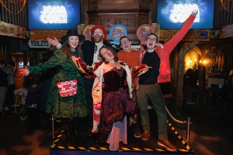 A group of five people is posing energetically on a small stage at an event called 'JONACON,' as indicated by the illuminated signs in the background. They are dressed in a mix of colourful and themed outfits, with some wearing hats, vibrant coats, and accessories. The person in the center is wearing a theatrical burgundy and pink dress with a patterned fur-lined coat, extending their arms dramatically. The others around them, wearing outfits including a green patterned coat, a red cap, and casual jackets, have wide smiles and raised arms. The setting appears to be an indoor event space with warm lighting, wooden decor, and a lively atmosphere, with other attendees visible in the background.