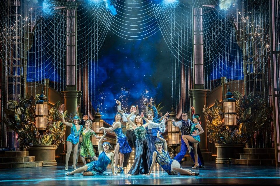 Amber Davies stands centre stage in a sparkling gold dress, surrounded by dancers in glamorous 1920s partywear, mid-routine during a Gatsby party scene.