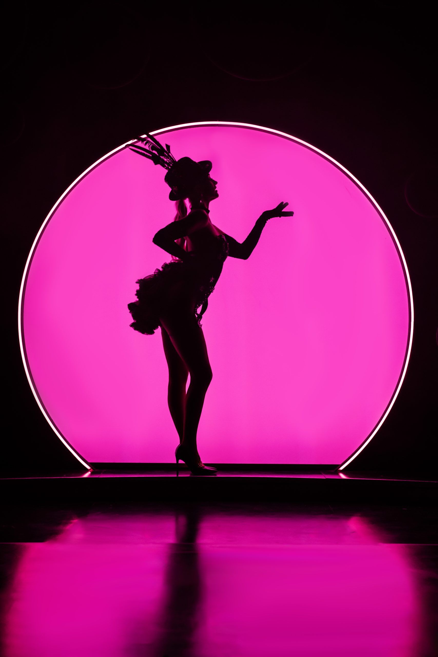 Silhouetted performer in heels and feathered headwear striking a classic burlesque pose in front of a vivid hot pink backlit circle, creating a striking contrast between figure and background.