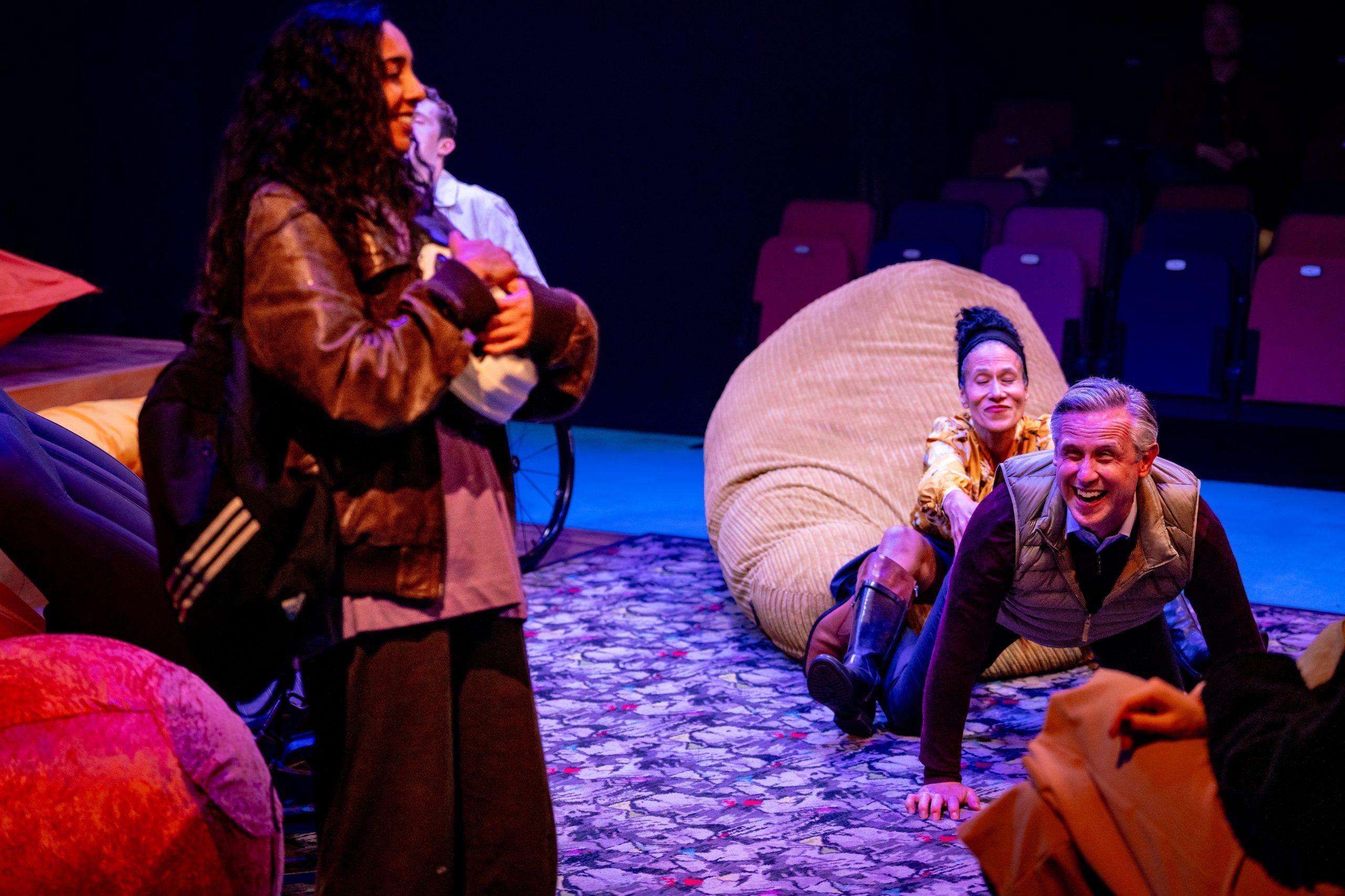 Jaz stands in the foreground holding a helmet, while Richard and Susan simulate sex on stage behind her