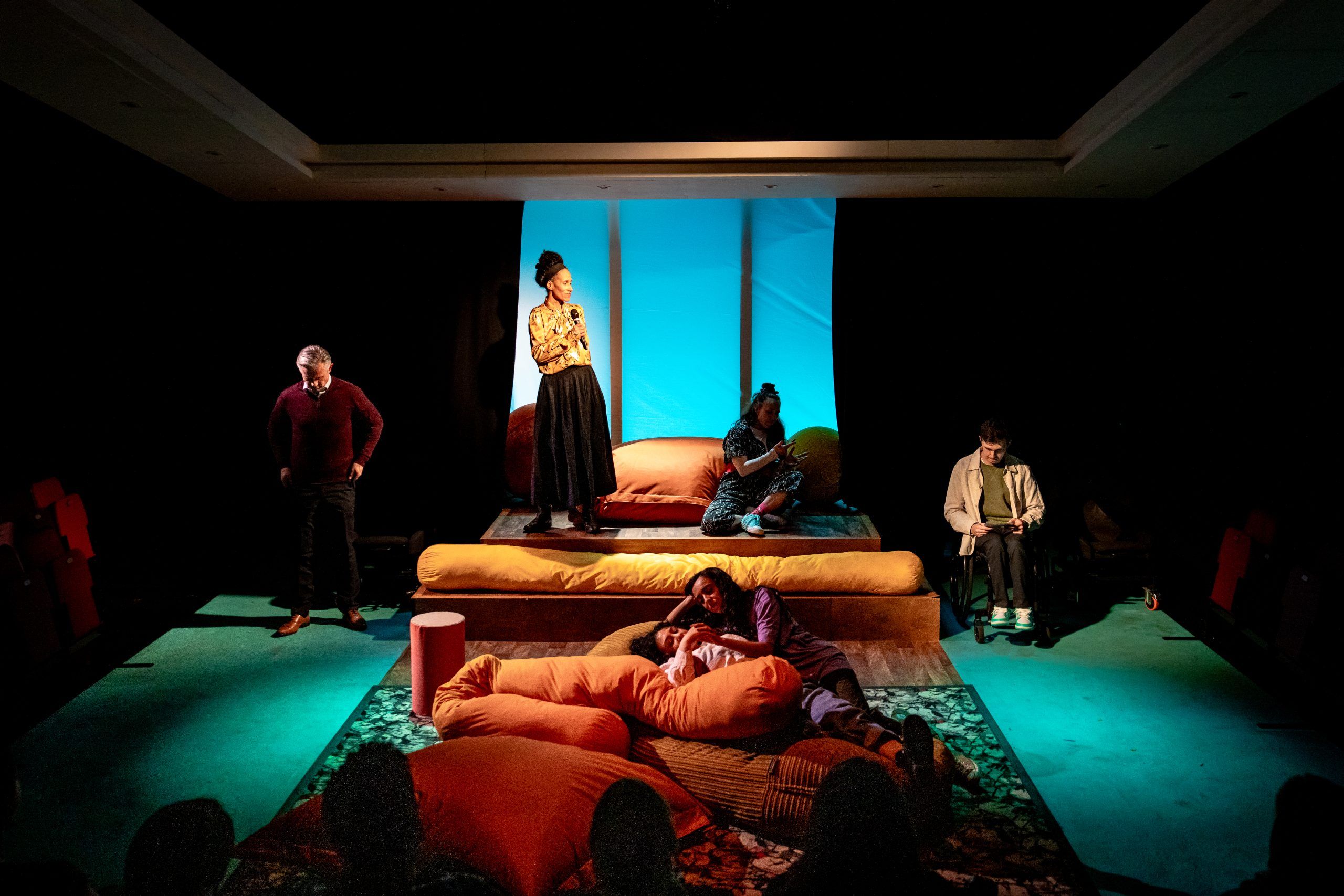 All six cast members spread across the stage in different zones, with warm blue lighting and oversized cushions