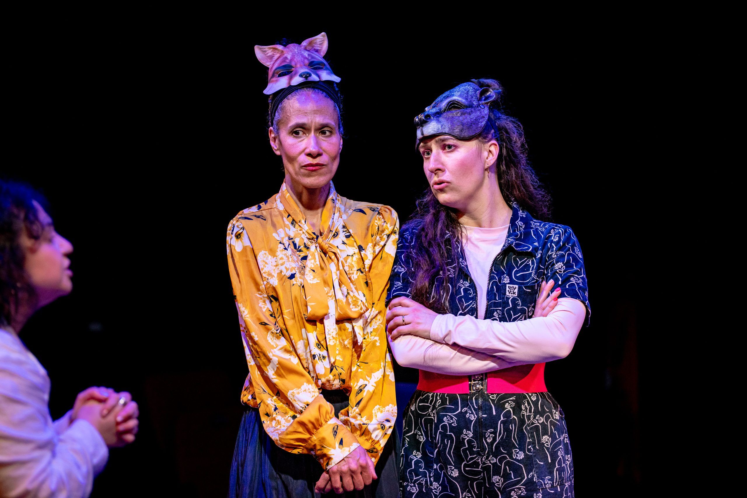 Susan and Sandra stand side by side wearing animal masks, arms crossed and faces serious, while Maya pleads with them