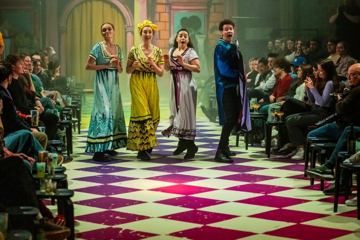 Four actors (Brigitte Freeme, Emma Andreatta, Monique Sallé, and Tim Walker) grouped together dramatically mid-scene, reacting theatrically to something behind camera, while audience members grin.