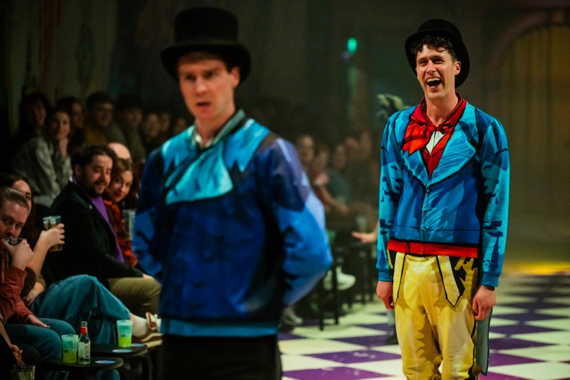 Andrew Macmillan laughs joyfully in the background wearing his cartoonish blue suit, as Tim Walker, in matching Regency-style outfit and top hat, stands in the foreground delivering a hilariously serious performance, the amused audience visible to the side.