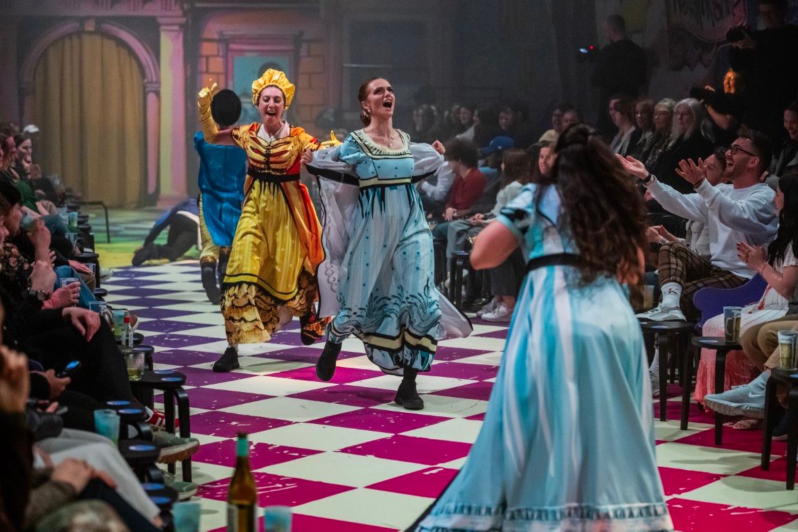 Brigitte Freeme (Lizzie) and Emma Andreatta (Mrs Bennet) enter dramatically from opposite sides – Lizzie with arms outstretched, Mrs B in full theatrical flight – while Monique Sallé (Jane) approaches from the other direction, seen from behind.