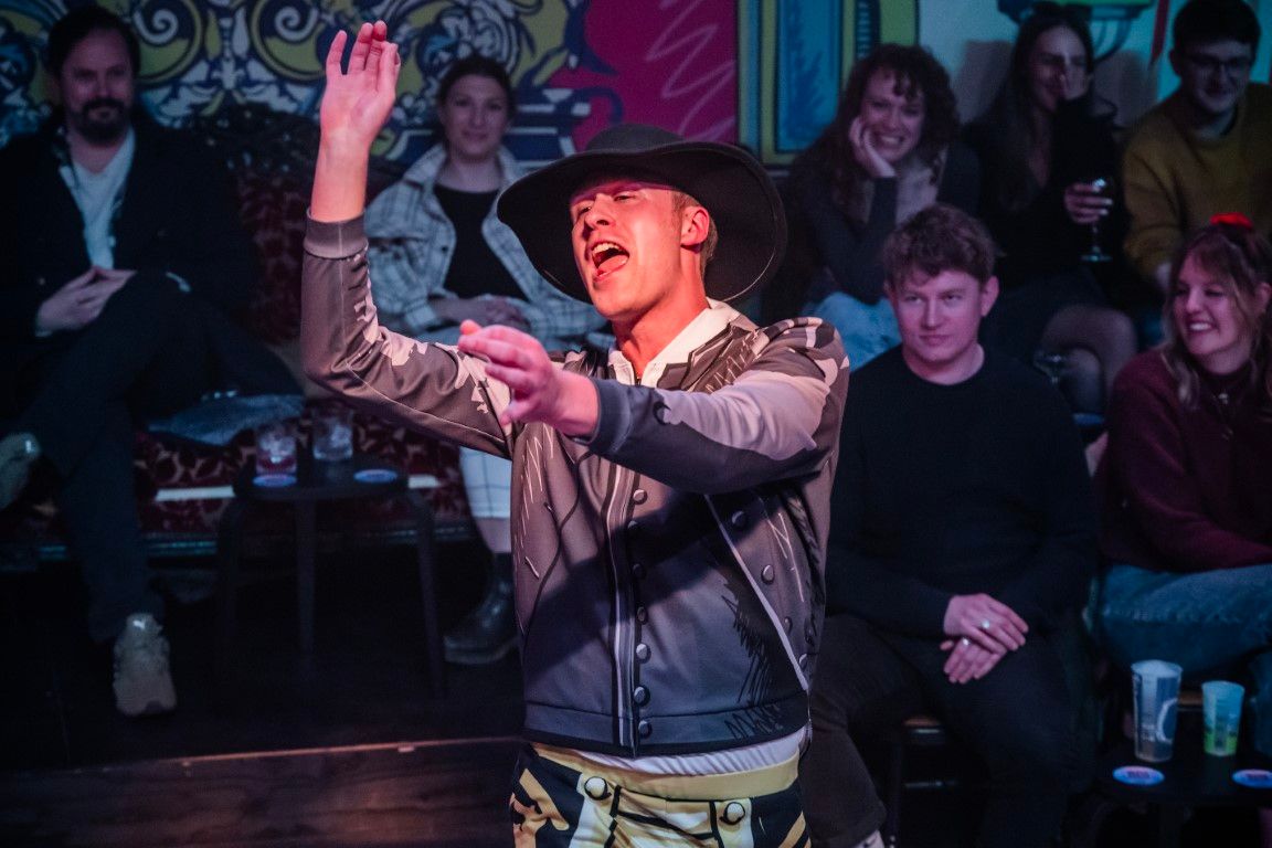Andrew Macmillan as Mr Collins, mid-serenade, dramatically gesturing in a grey cartoon-printed suit and wide-brimmed hat, hamming it up to a bemused audience.