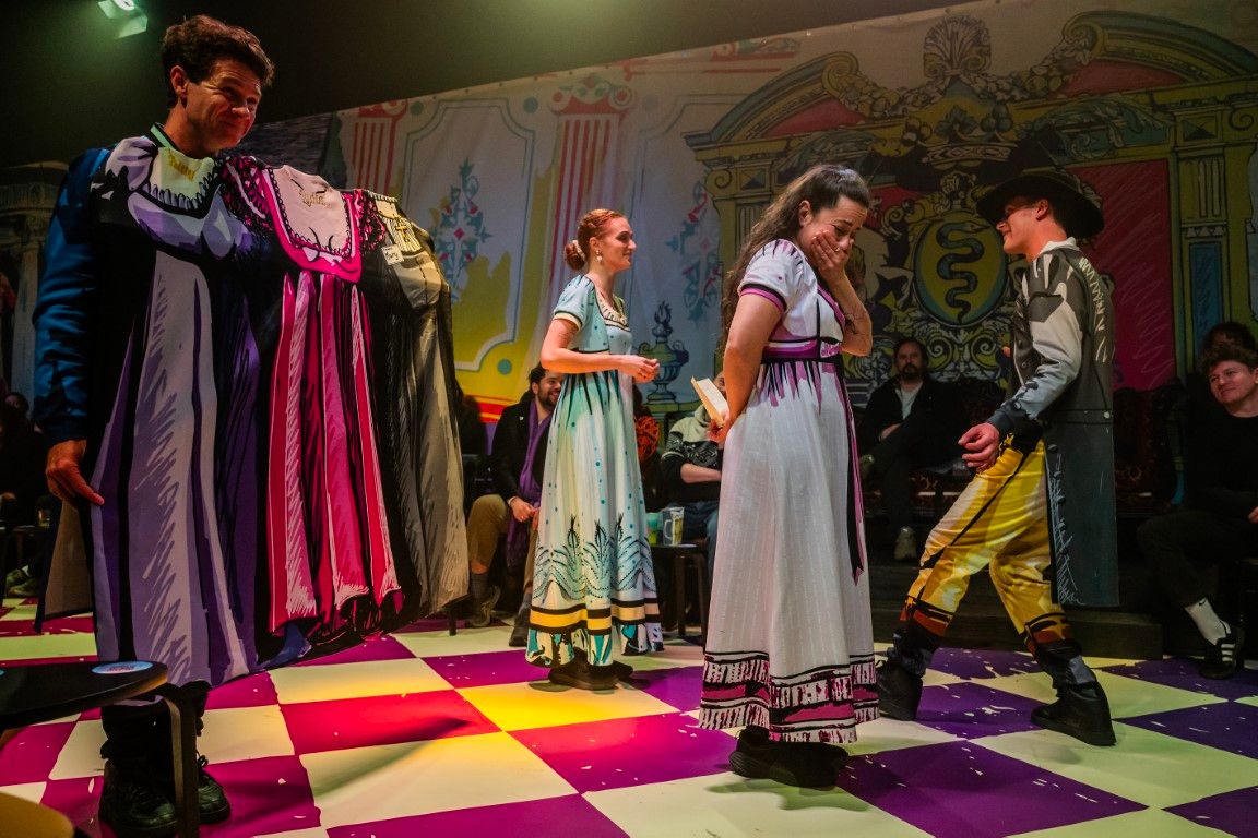 Tim Walker comically holds up multiple cartoon-style dresses attached to his costume, while cast members Monique Sallé, Brigitte Freeme, and Andrew Macmillan laugh at the silliness.