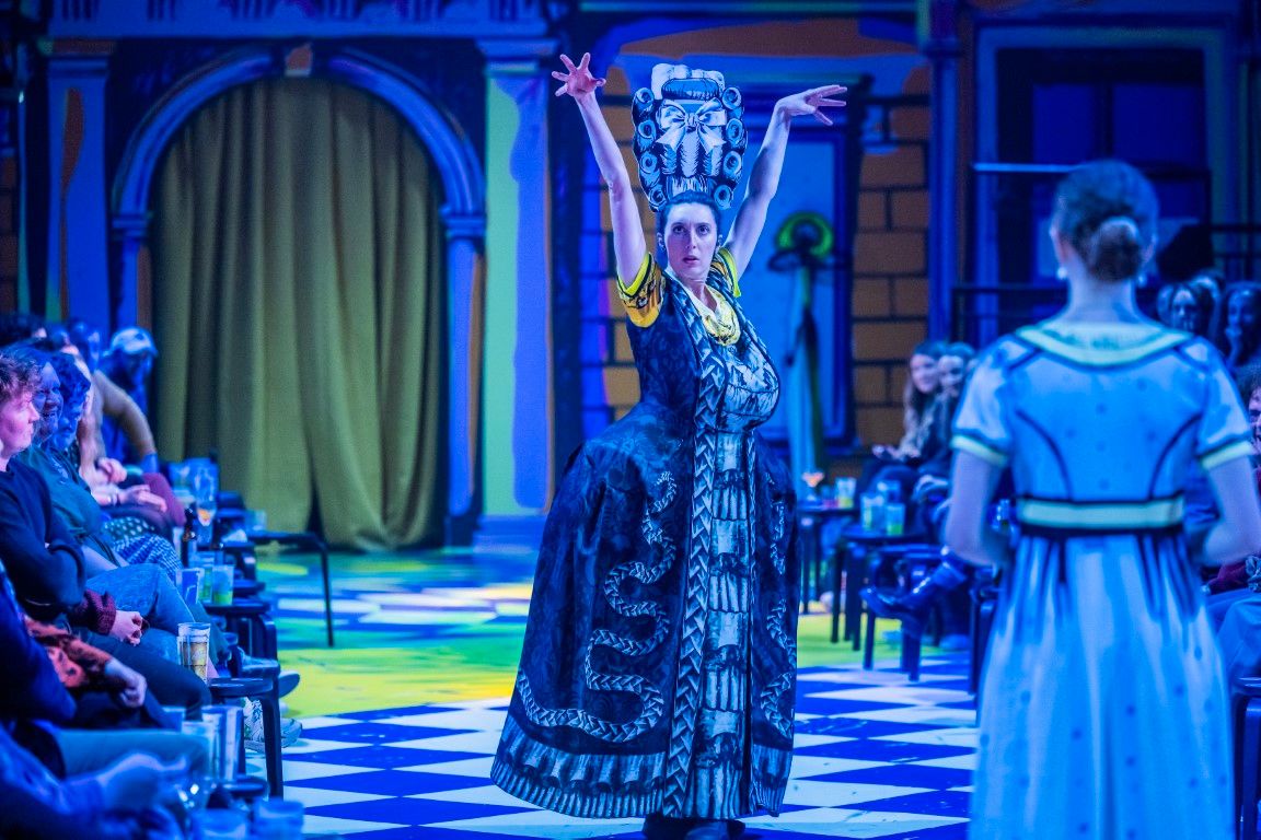 Emma Andreatta (Lady Catherine de Bourgh) strikes a bold, operatic pose centre stage in an elaborate printed costume that mimics period design, complete with a towering cartoonish wig. The set, bathed in moody blue light, surrounds her with oversized illustrated arches and checkerboard flooring, while the audience watches on from both sides.