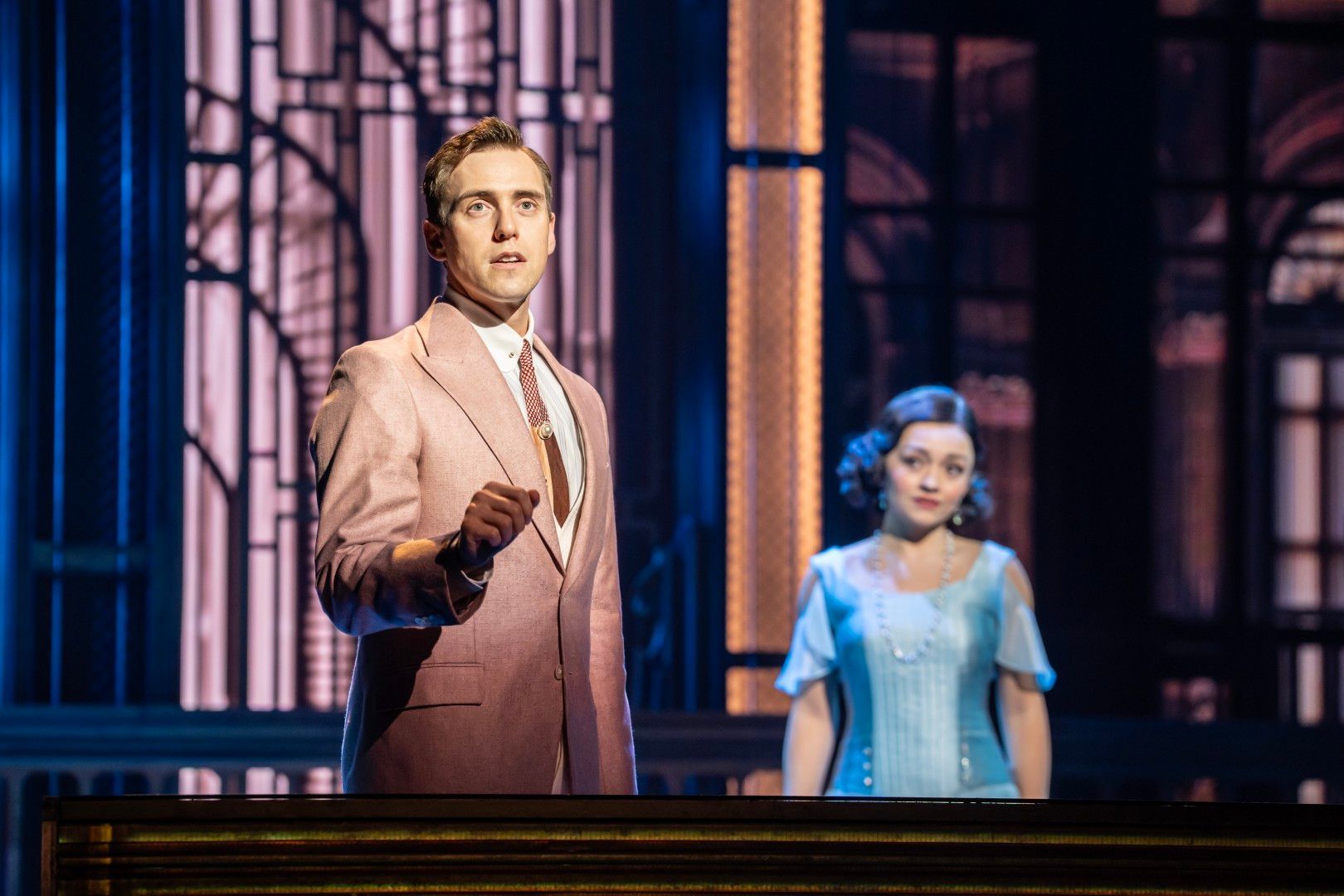 Jamie Muscato as Gatsby stands mid-song in a pink suit, with Frances Mayli McCann as Daisy watching him quietly in the background.