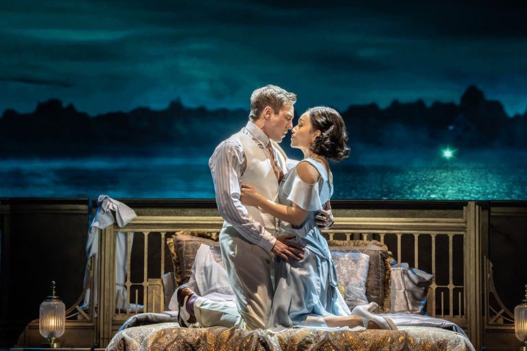 Jamie Muscato and Frances Mayli McCann embrace during a poignant duet, standing before a romantic seascape backdrop that blends digital projection with real set elements.