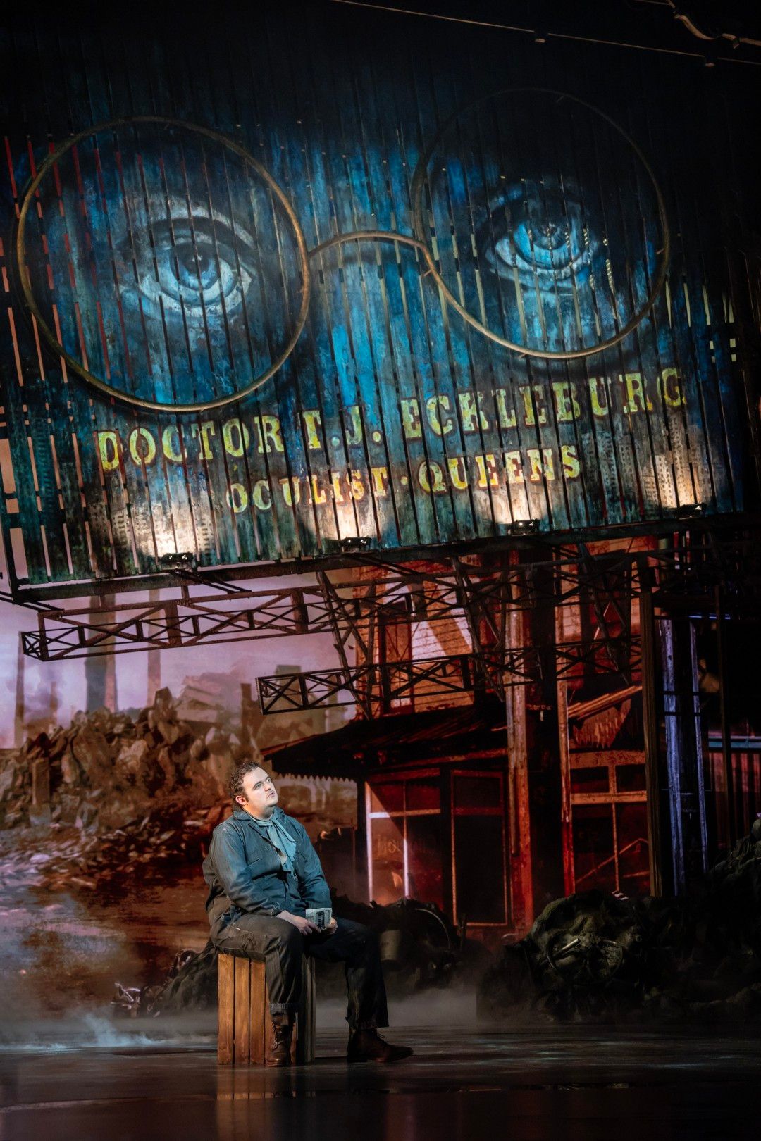 Joel Montague as George Wilson stands beside a vintage petrol pump, mid-song, with a weathered garage set behind him and a neon optician sign glowing faintly.