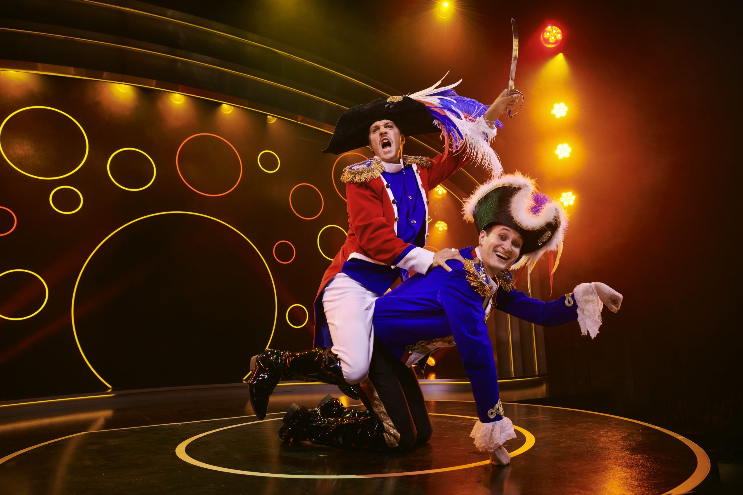 Rémi Martin and Spencer Novich in flamboyant Napoleonic costumes mid-performance. Rémi is perched on Spencer's back, while he's on all fours grinning up at the audience under warm theatrical lights.