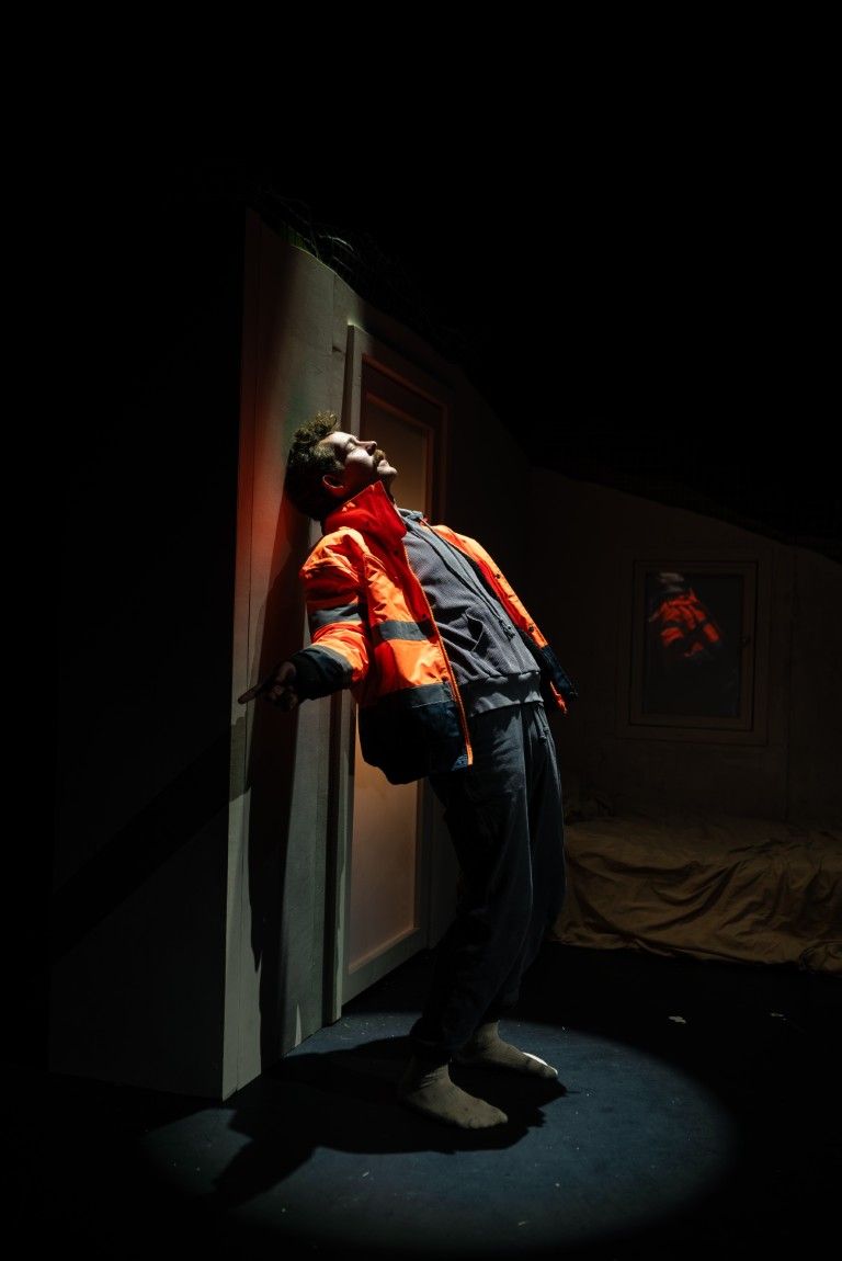 A man leans against a wall under a spotlight, wearing a bright orange jacket, his body language full of tension and struggle.