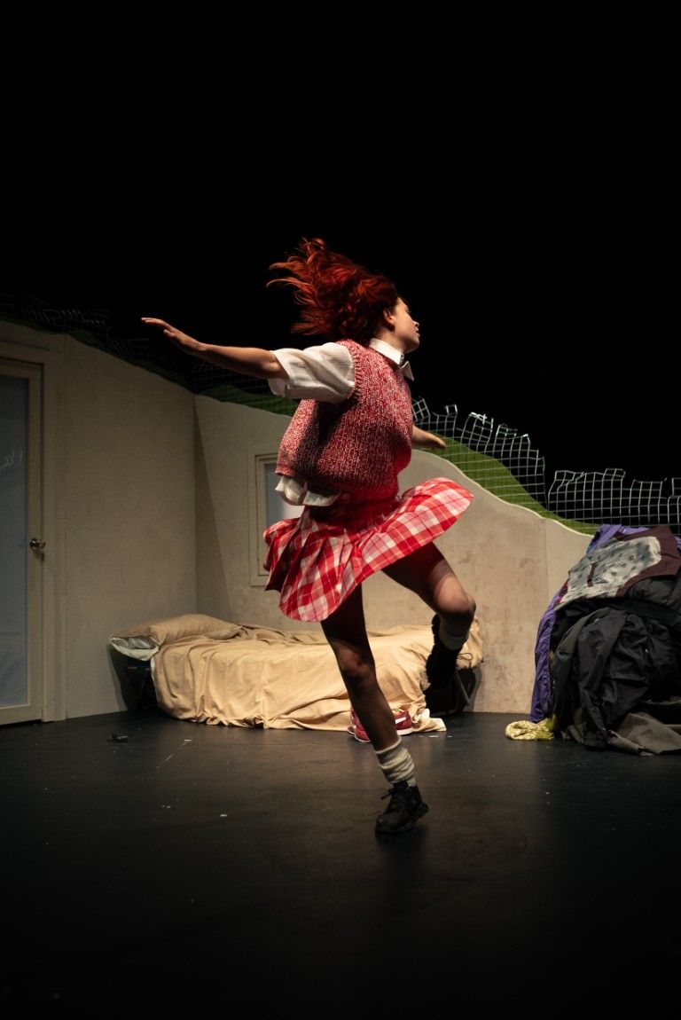 A young woman in a red checked skirt leaps mid-dance across a sparse bedroom set, her hair flying, full of energy and freedom.