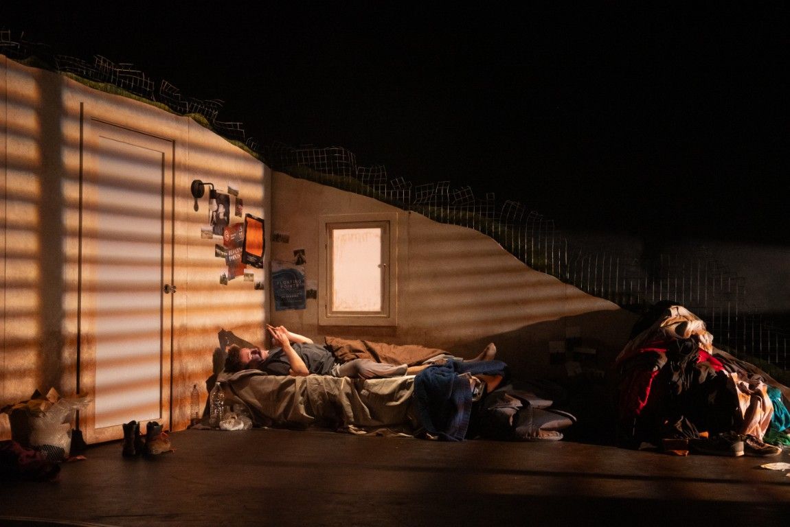 A messy bedroom set, strewn with clothes and clutter. A man lies on a bed scrolling through his phone, lit by warm striped shadows.