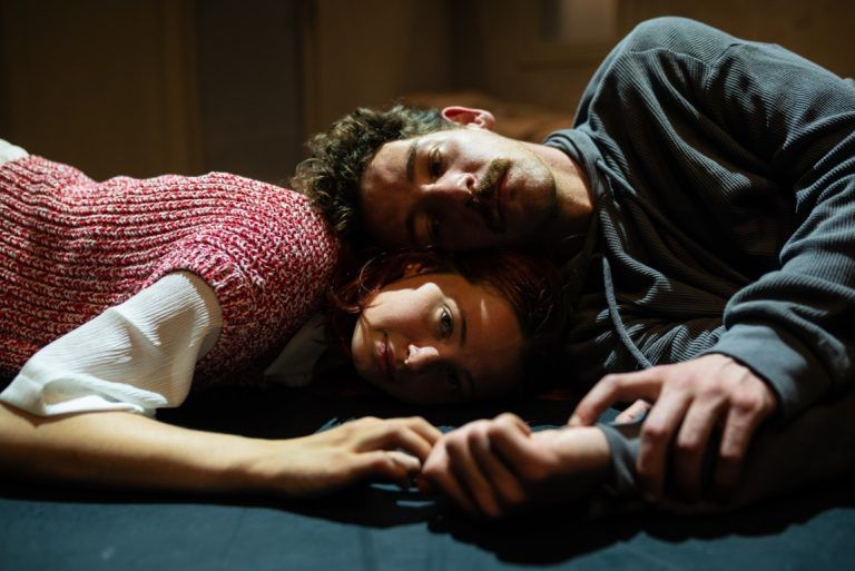 A young man and a woman lie on the floor, their heads resting together, both looking lost in thought. Their hands are loosely intertwined.