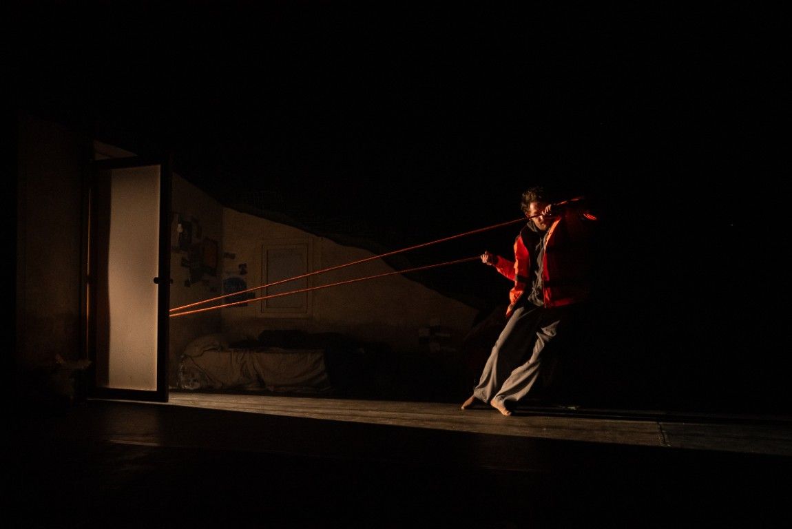 A man pulls against red cords stretched taut across a dark stage, fighting to move forward. A glowing doorway looms behind him.