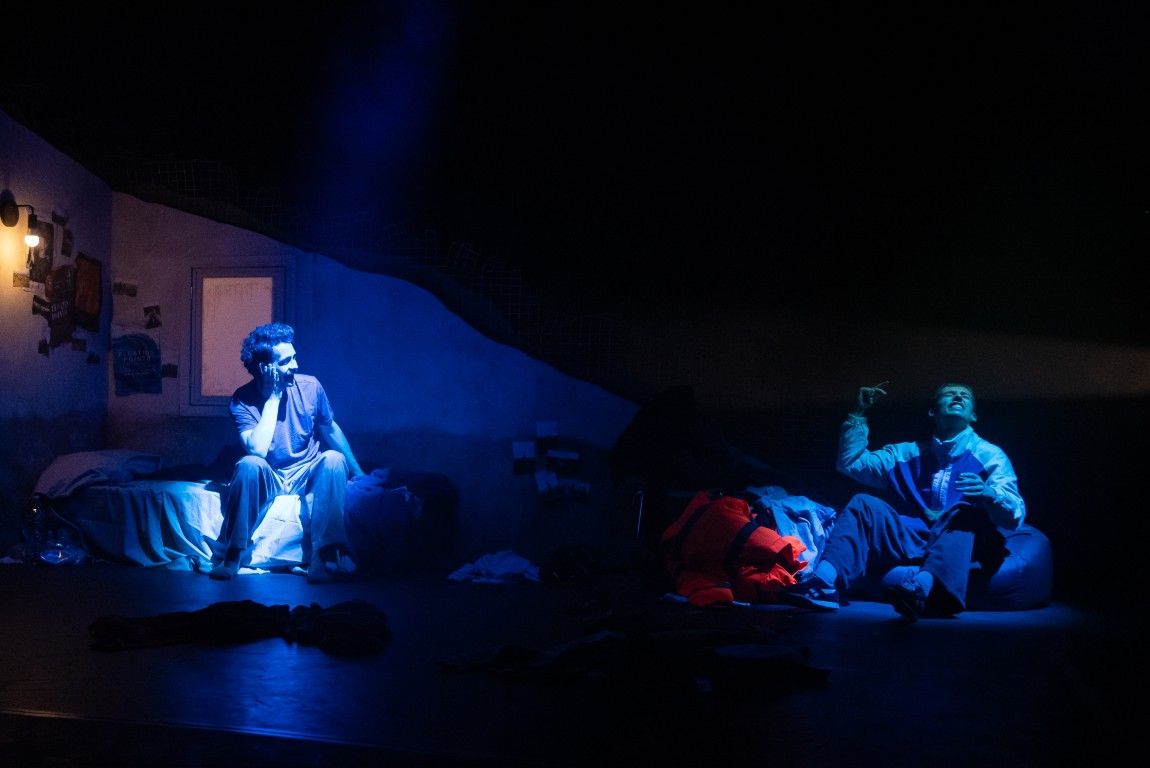 Two young men sit separately in the same dark bedroom. One is lit in blue on the bed, the other animatedly talks on the floor.