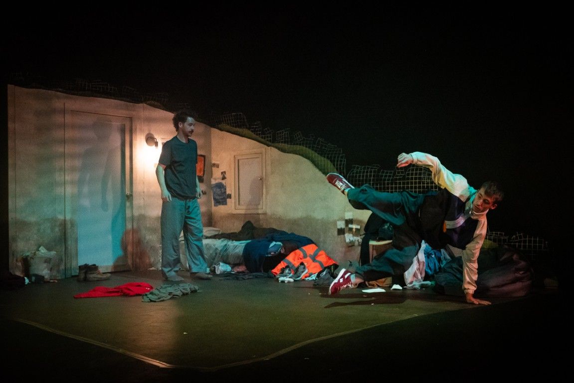 Two young men in a sparse bedroom set. One stands watching, while the other kicks out dramatically mid-dance, surrounded by piles of clothes.