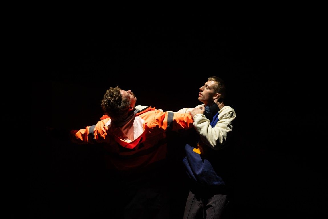 Two young men caught in a powerful moment, one pulling the other close under stark lighting. One wears a bright orange work jacket.
