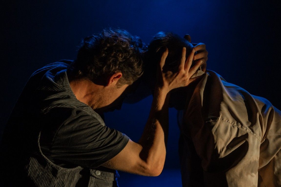 Two men press their foreheads together, hands cradling each other’s heads in a gesture of connection, grief, and silent support.