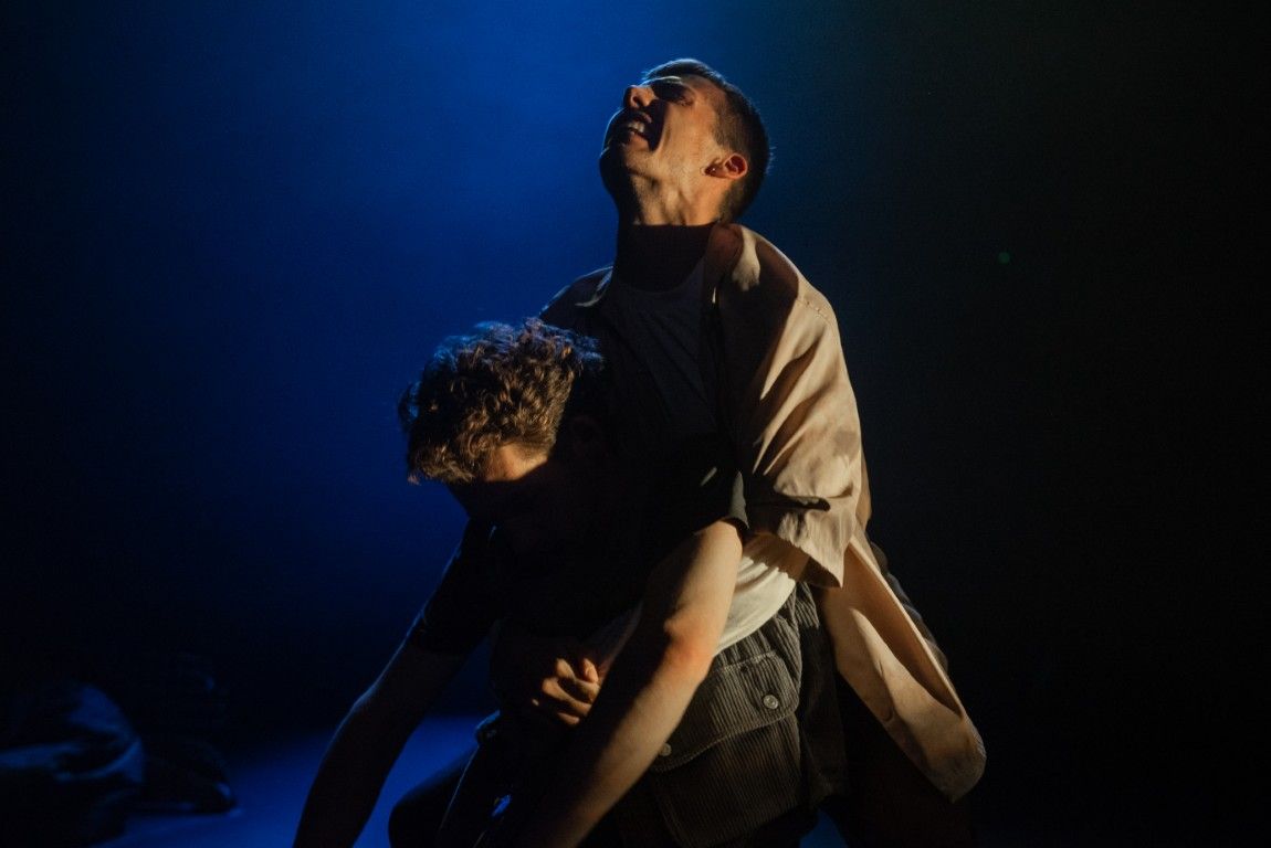 A raw, emotional moment as one man collapses into another’s arms. One man looks upwards, his face contorted in pain under blue light.