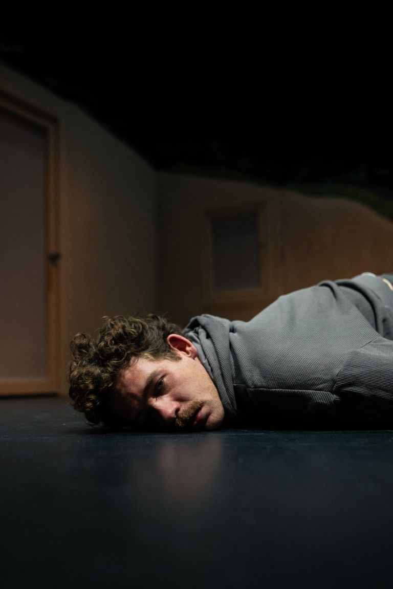 A young man lies face-down on the floor, staring ahead with a look of exhaustion and quiet sadness.